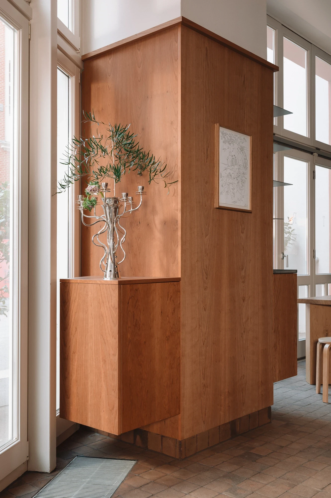 Strom Berlin by Celeste Asfour, photographed by Clemens Poloczek — A polished silver candelabra holds an olive branch on top of a cherry wood service column at the cafe's entrance beside a tall window.