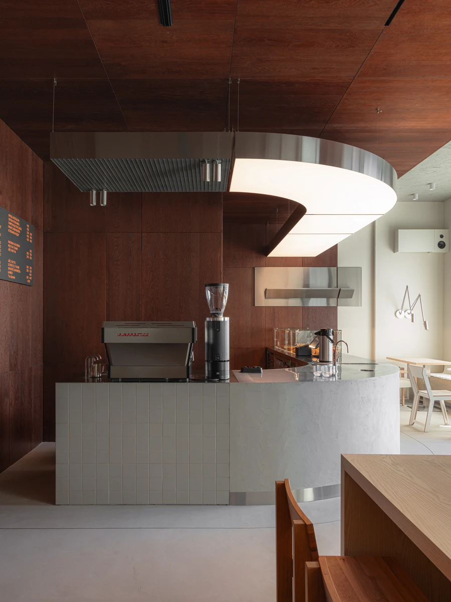 Biblioteka Coffee by Kidz, photographed by Alexander Plyaskin — the curved counter seen from the side, the espresso machine in the foreground and the contrast between dark oak walls and a pale poured floor reading as the project's central gesture.