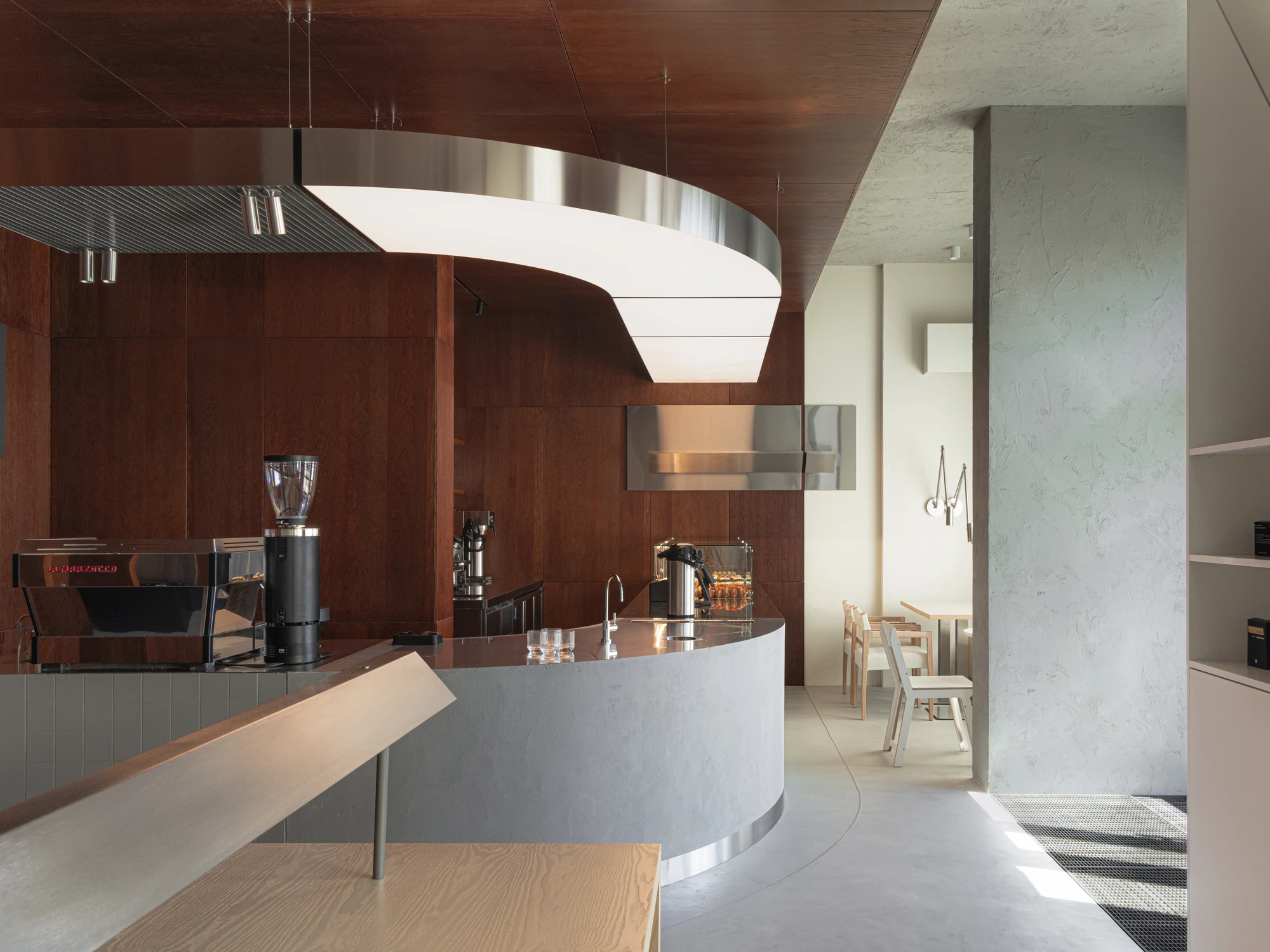 Biblioteka Coffee by Kidz, photographed by Alexander Plyaskin — a wide view of the cafe interior from the silence side, the curved bar and steel canopy visible at left, the polished plaster wall and an articulated brass lamp at right.