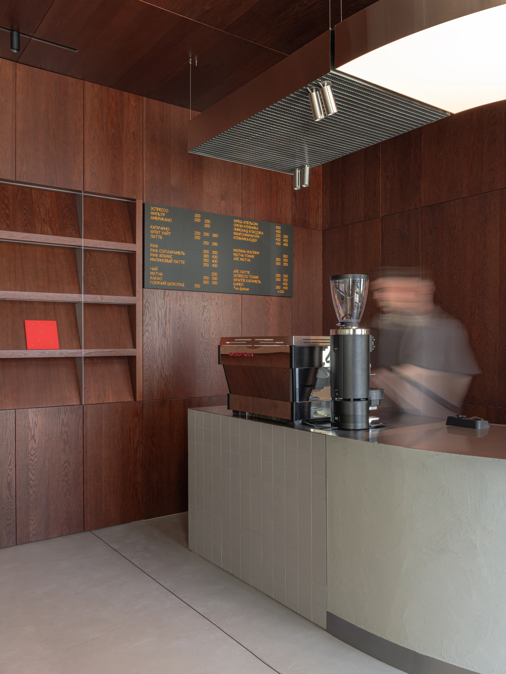 Biblioteka Coffee by Kidz, photographed by Alexander Plyaskin — a dark oak panelled wall holds the printed menu in cyrillic above a microcement counter, with a barista visible only as motion blur passing behind the espresso machine.