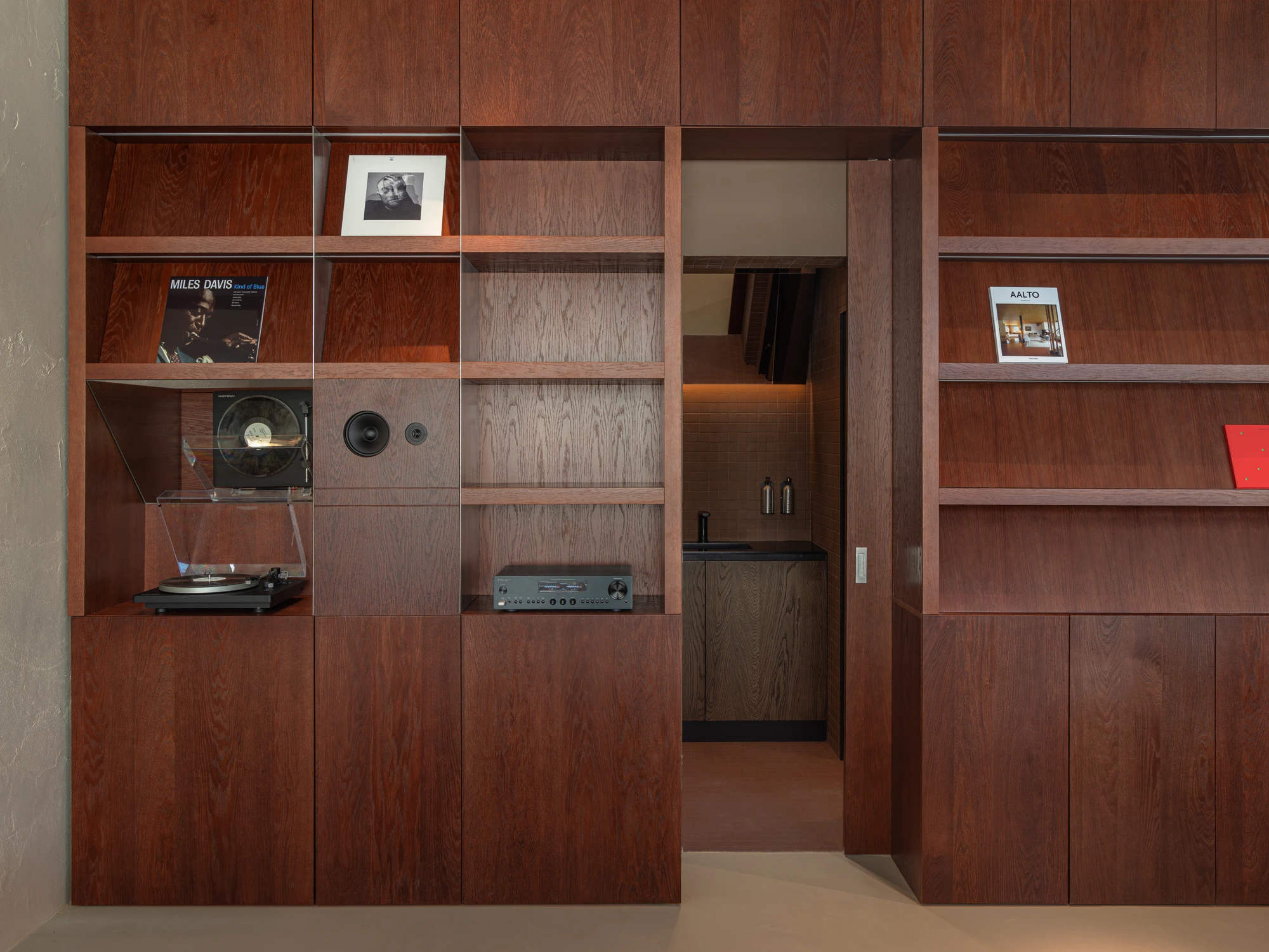 Biblioteka Coffee by Kidz, photographed by Alexander Plyaskin — the full-height dark oak shelving wall with vinyl, a turntable and an amplifier on the lower shelf and a hidden sliding door opened to reveal a small back corridor.