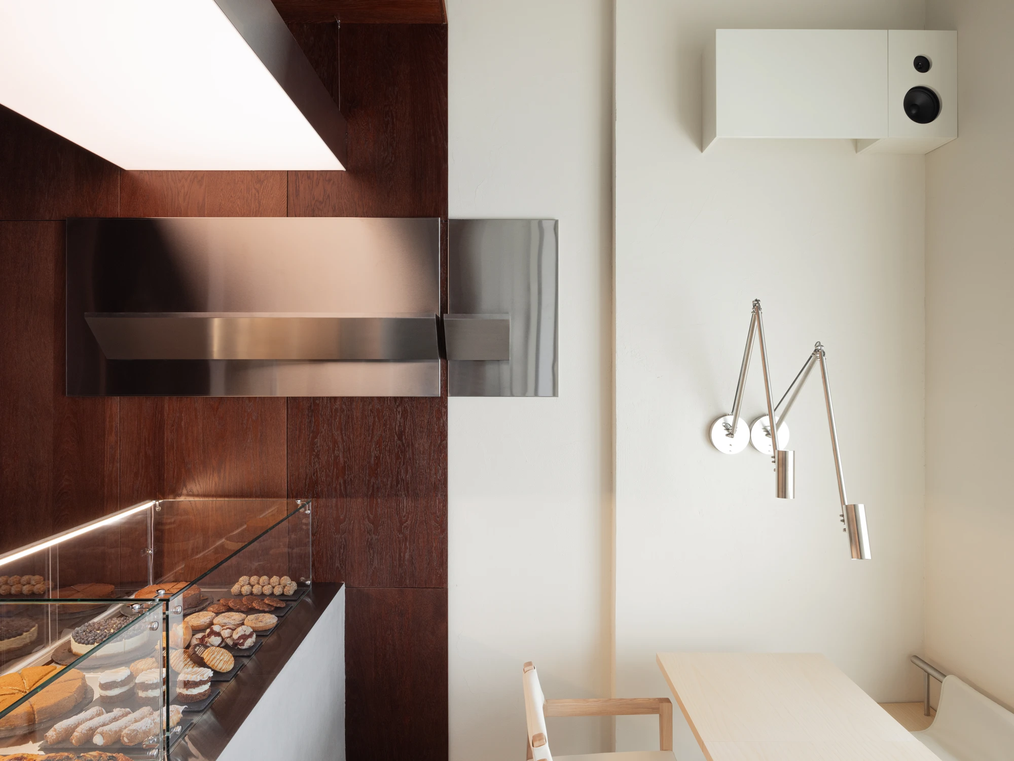 Biblioteka Coffee by Kidz, photographed by Alexander Plyaskin — a pastry display case in polished steel set into the dark oak wall, with the articulated brass lamp and a corner of the small bistro table visible at right.