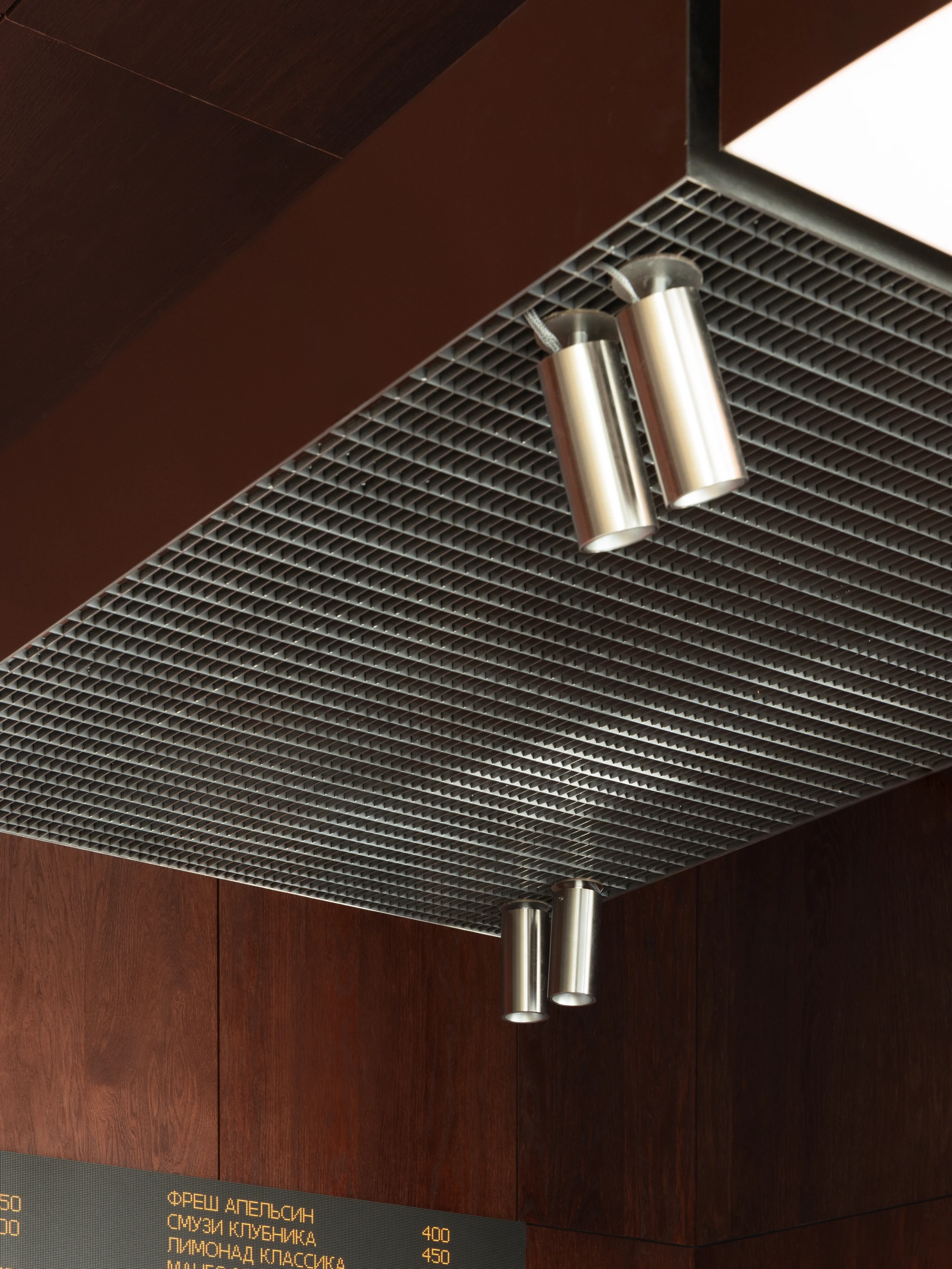 Biblioteka Coffee by Kidz, photographed by Alexander Plyaskin — an overhead detail of the mud-grate ceiling element above the espresso machine, two cylindrical track lamps angled across the fine metal grille.