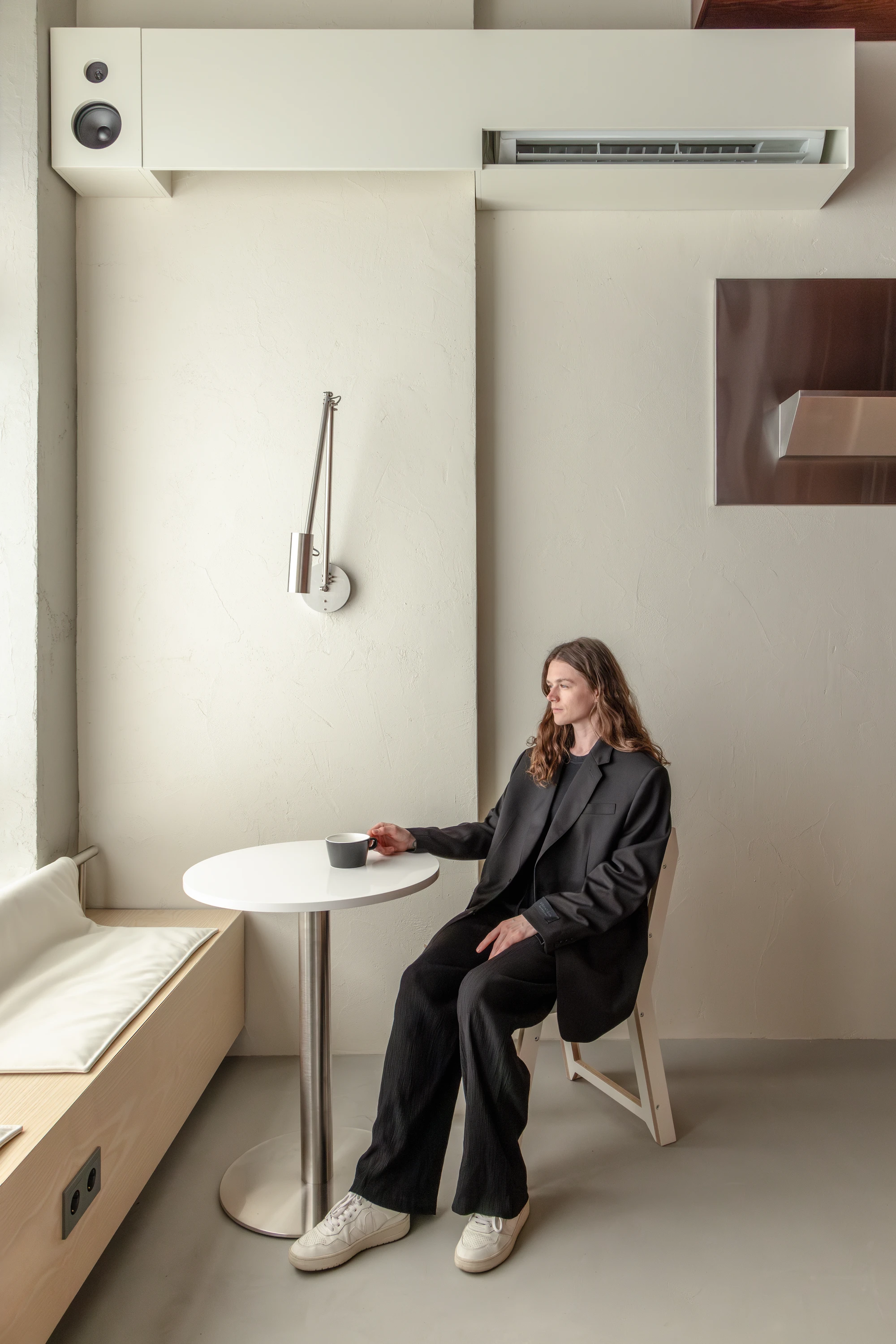 Biblioteka Coffee by Kidz, photographed by Alexander Plyaskin — a woman in a black coat sits at a small round bistro table beside the panoramic window, an articulated brass wall lamp mounted above her on the plaster wall.
