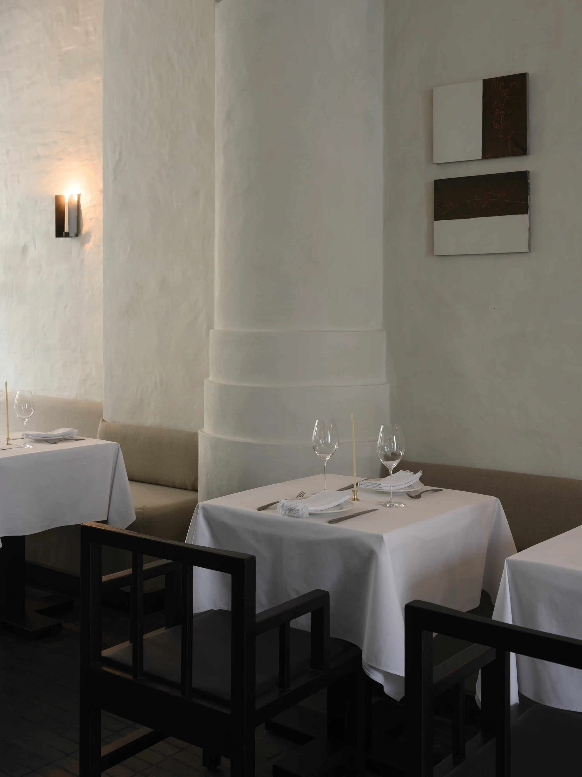 OKN Restaurant by SAGA, photographed by Daniel Annenkov — A plaster column in the main dining room, set beside two white-linen tables with brass taper candles. Two rectangular geometric artworks hang on the plaster wall at right.