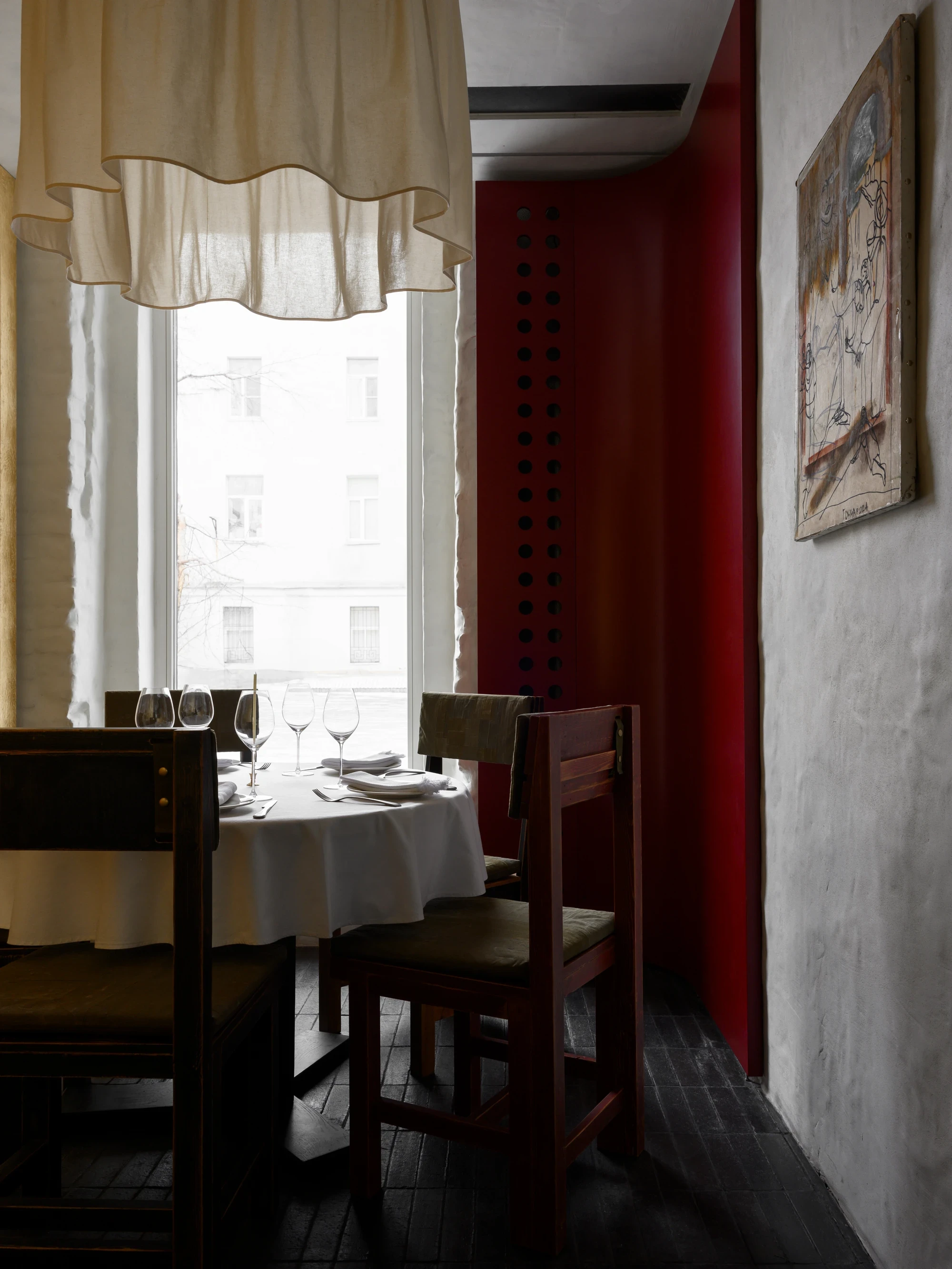 OKN Restaurant by SAGA, photographed by Daniel Annenkov — A window table in the red-room zone: dark timber chairs with canvas upholstery, a round table with white linen, cream curtains with a scalloped hem and a deep crimson wall at right.