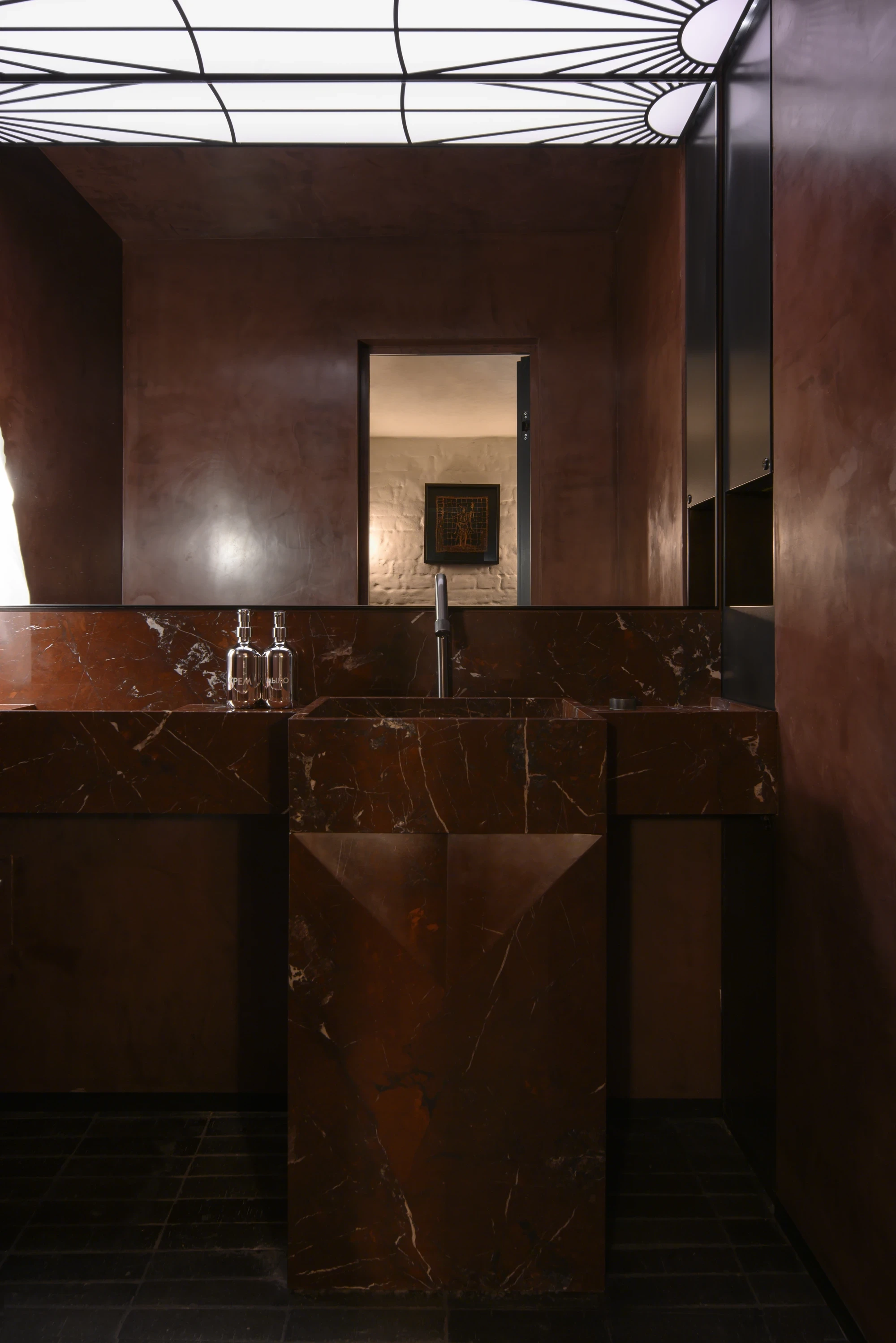 OKN Restaurant by SAGA, photographed by Daniel Annenkov — The bathroom vanity: a monolithic unit of deep brown-veined marble with a faceted geometric pedestal sink. A backlit panel with a sunburst spoke motif in black and white provides the room's sole li