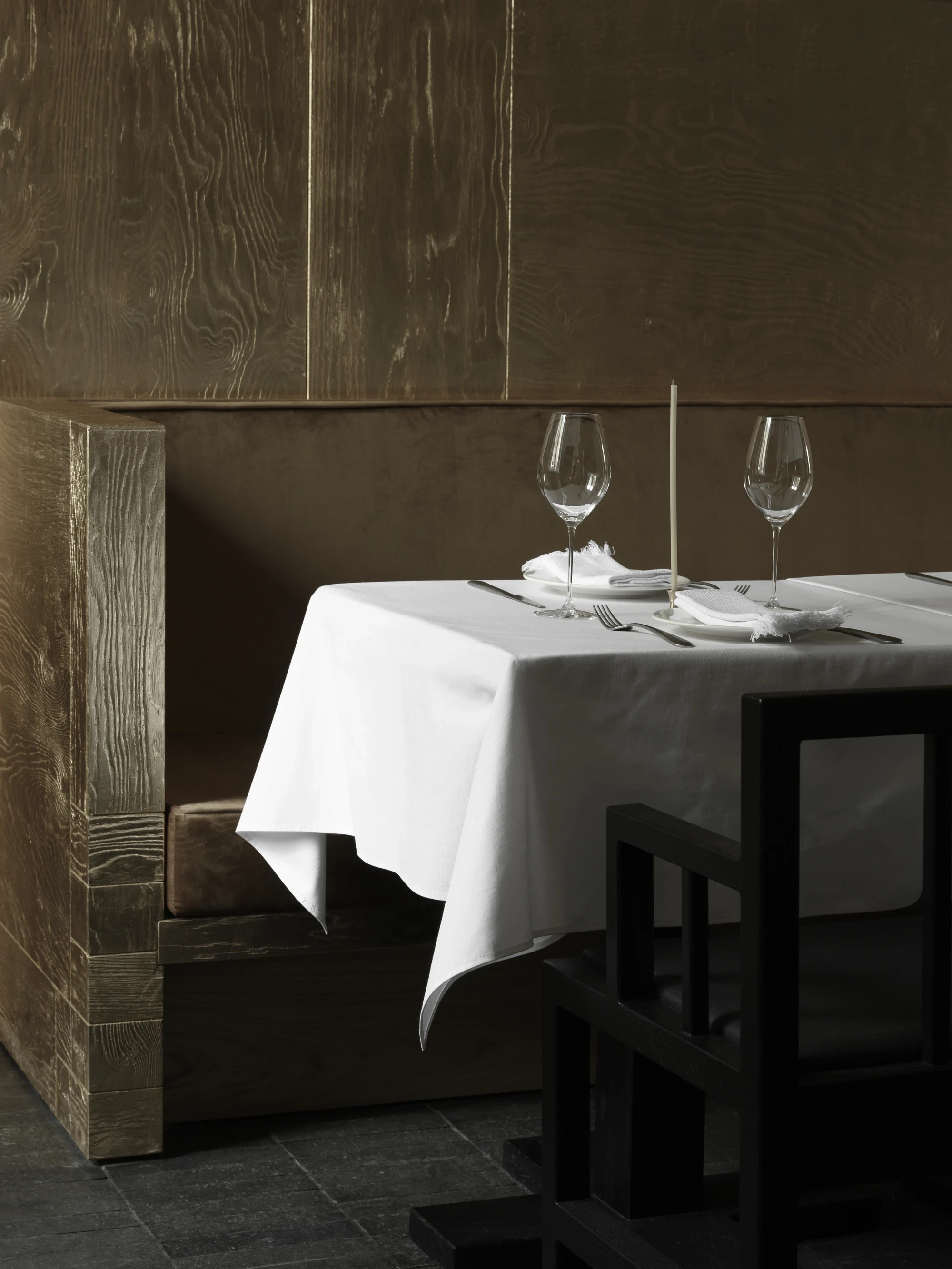 OKN Restaurant by SAGA, photographed by Daniel Annenkov — A banquette table set for two, the white linen cloth bright against the matte gold-leaf timber panels behind. Two wine glasses and a single taper candle are the only objects on the table.