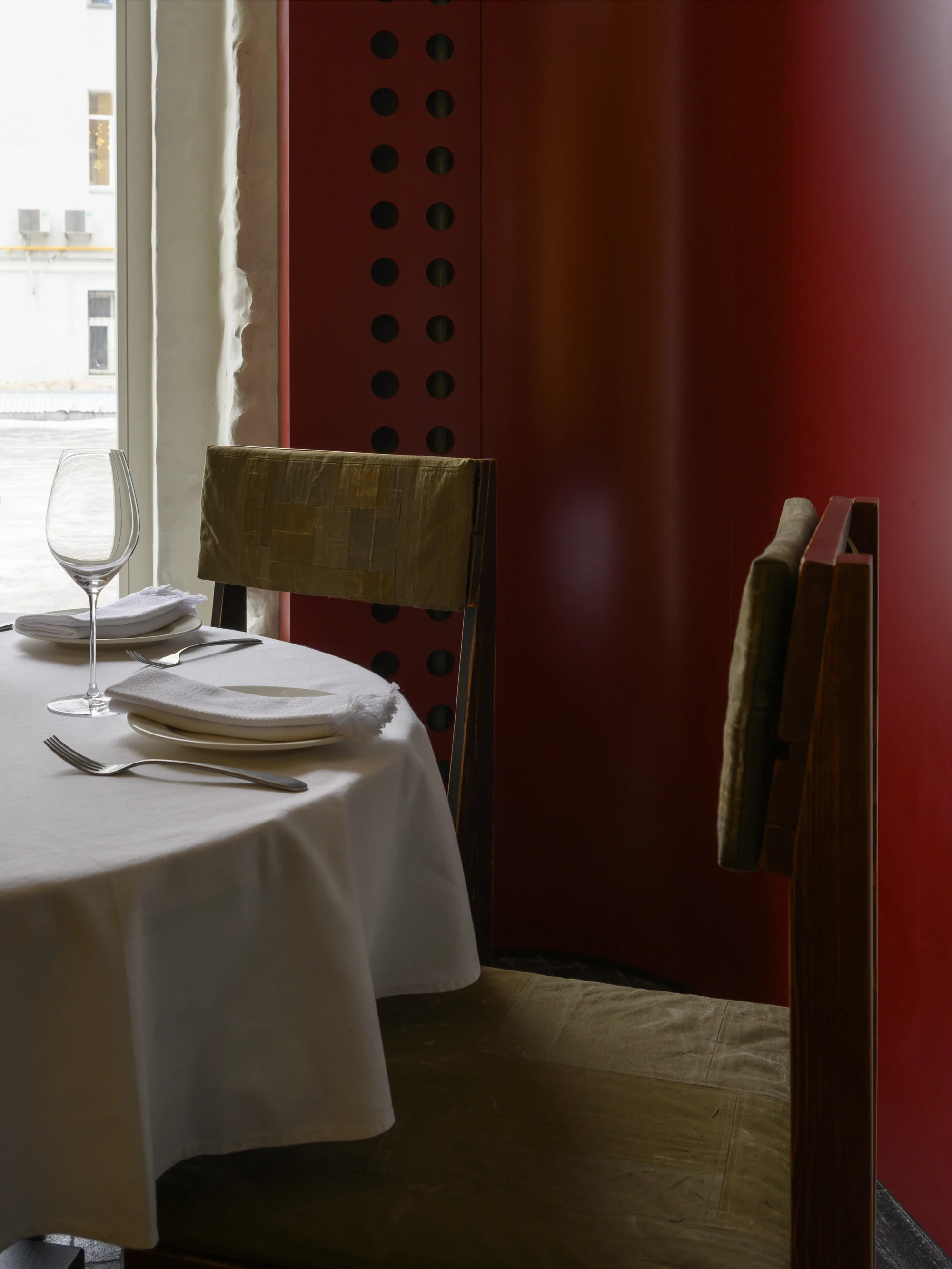 OKN Restaurant by SAGA, photographed by Daniel Annenkov — Detail of the red-room seating area: a dark timber chair with olive canvas back leans against the deep crimson plaster wall, its grid of circular perforations visible.