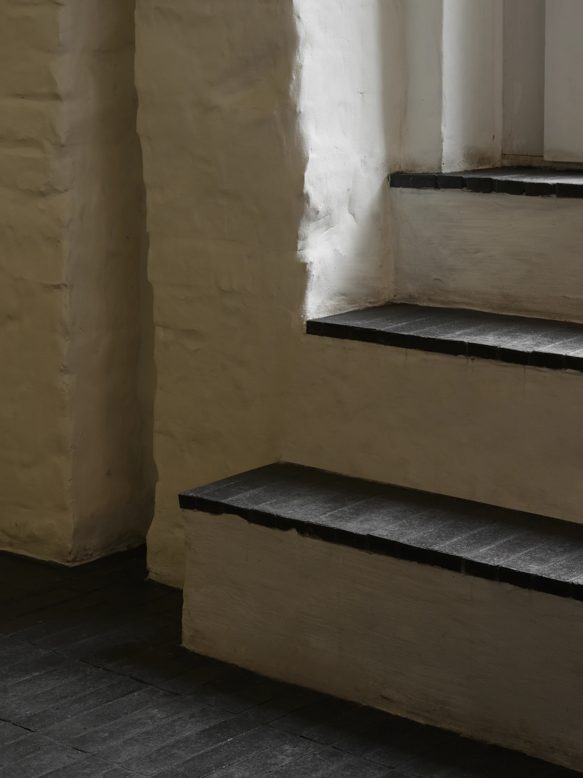 OKN Restaurant by SAGA, photographed by Daniel Annenkov — A tight corner of rough white plaster wall and black-painted timber window frame, with two slate-coloured stone steps rising to a lower window opening.