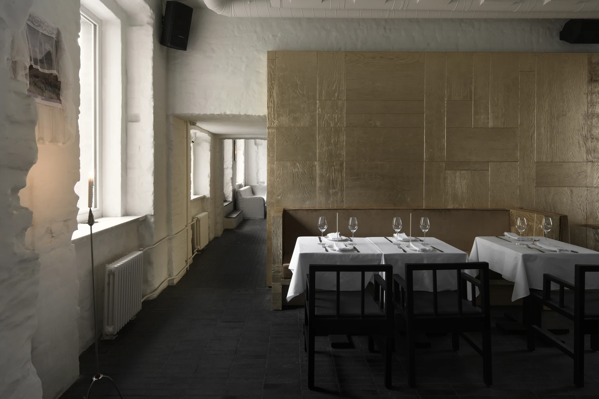 OKN Restaurant by SAGA, photographed by Daniel Annenkov — Long view along the main dining room in daylight: rough white plaster walls at left with tall windows, gold-leaf banquette partition running along the right. White-linen tables recede into depth.