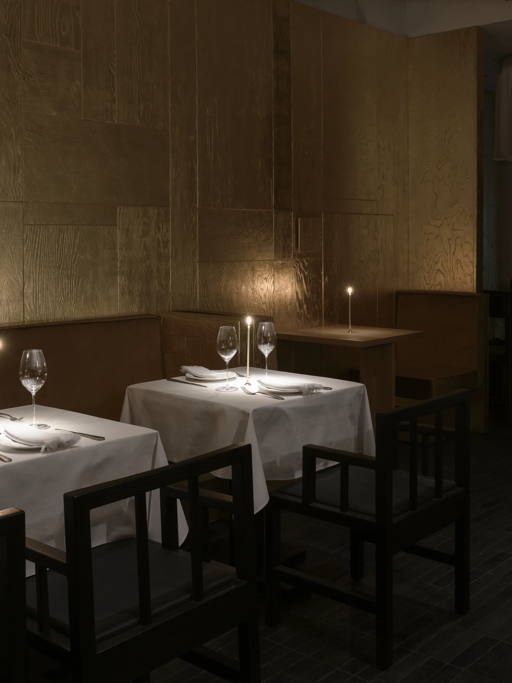 OKN Restaurant by SAGA, photographed by Daniel Annenkov — Candlelit evening view of the banquette dining area: brass taper candles on white-linen tables cast pools of warm light onto the gold-leaf timber wall panels, which glow amber.