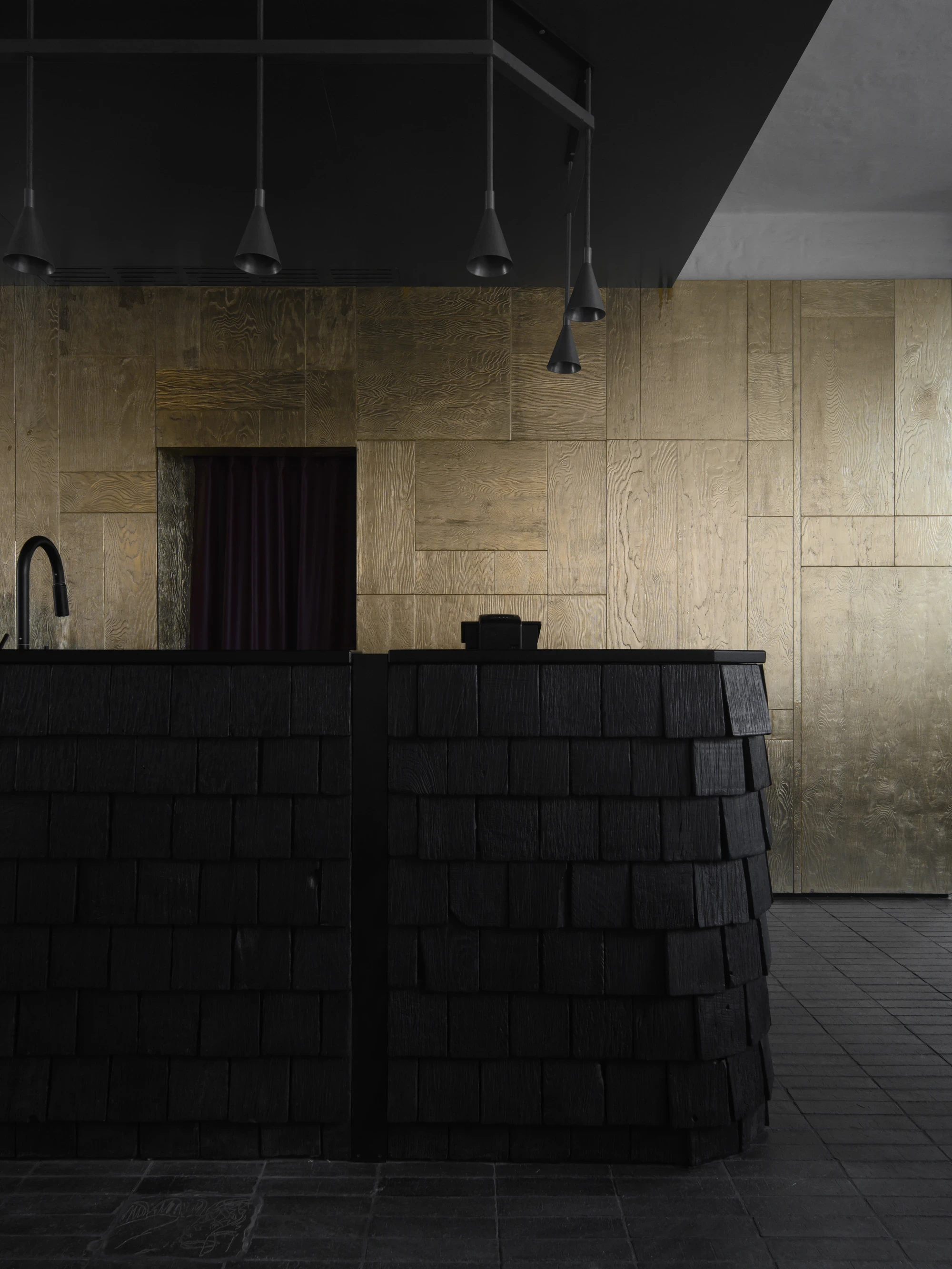 OKN Restaurant by SAGA, photographed by Daniel Annenkov — The bar counter of OKN Restaurant, clad in horizontal courses of black-charred timber shingles, stands before a wall of wood-grained gold-leaf panels beneath a row of black pendant lights.