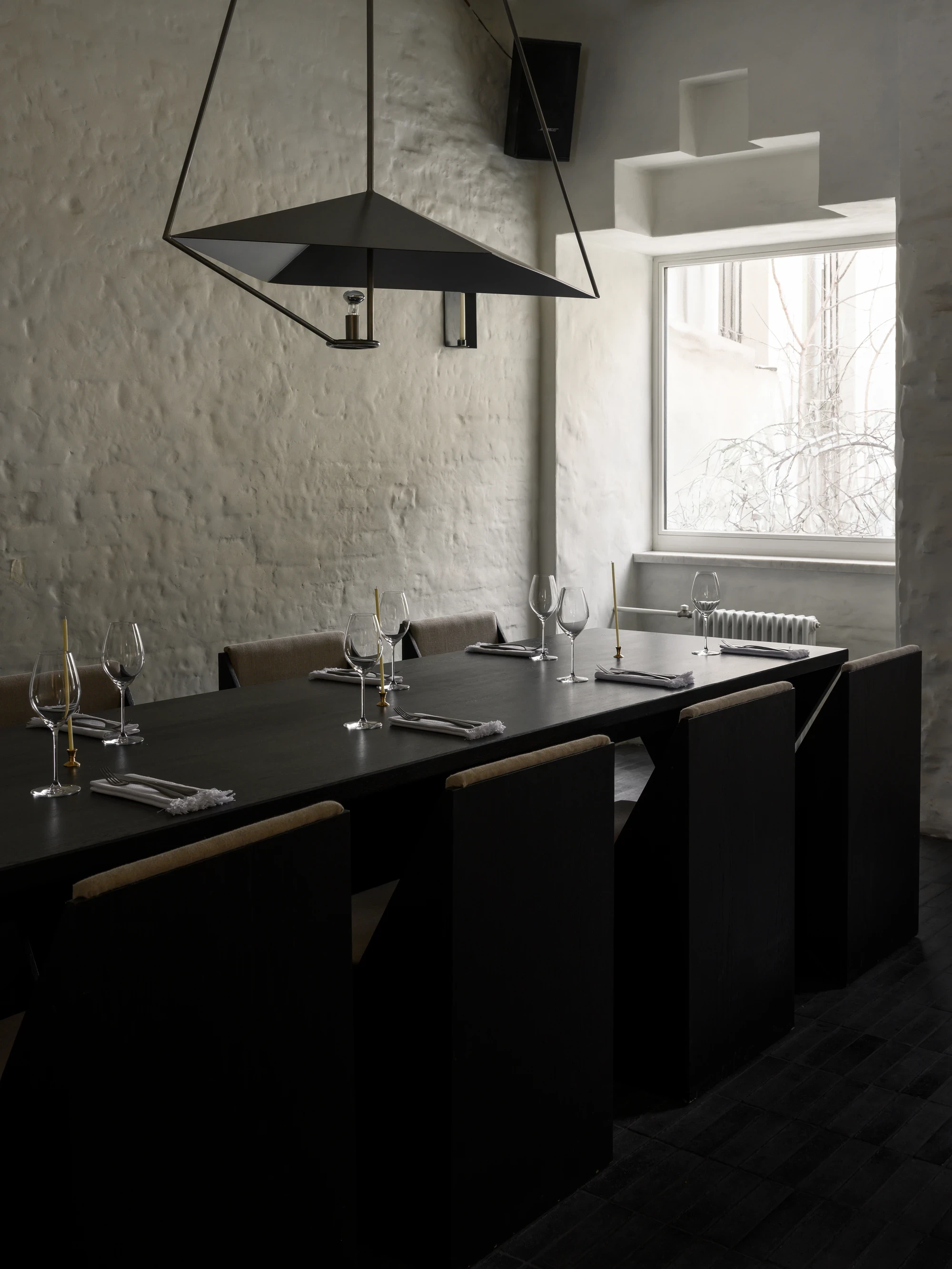 OKN Restaurant by SAGA, photographed by Daniel Annenkov — The private dining room with a black matte geometric pendant light suspended over a long dark table. Black upholstered chairs with fabric backs wrap the table against rough white plaster walls.