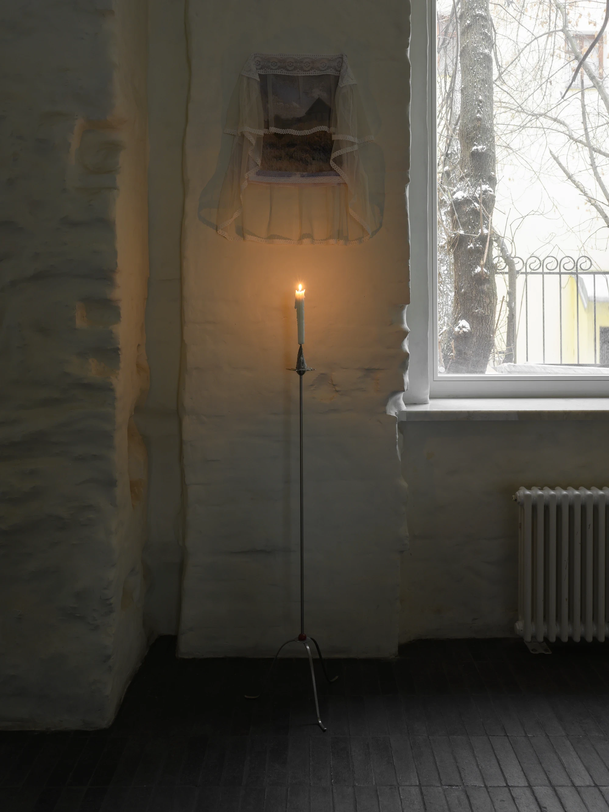 OKN Restaurant by SAGA, photographed by Daniel Annenkov — A tall tripod steel candle-stand bearing a lit white taper candle, positioned in front of rough textured white plaster wall. A small textile artwork is mounted in the deep window embrasure above.