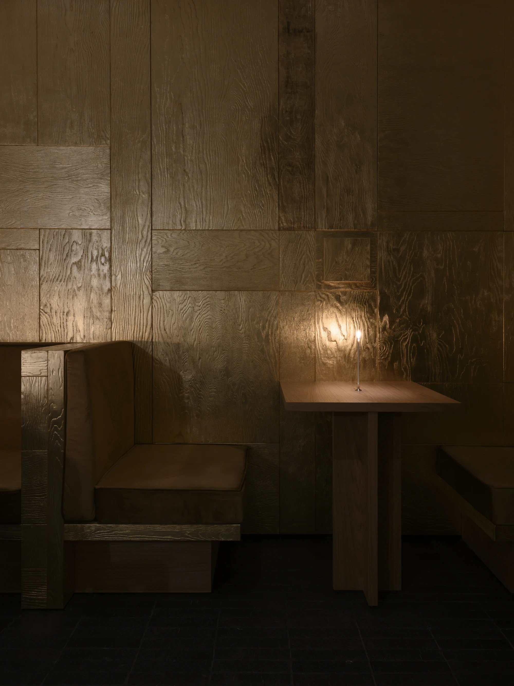 OKN Restaurant by SAGA, photographed by Daniel Annenkov — Evening candlelight view of the banquette zone: a single brass taper candle on a side table glows against the gold-leaf timber wall panel, turning it amber-ochre.
