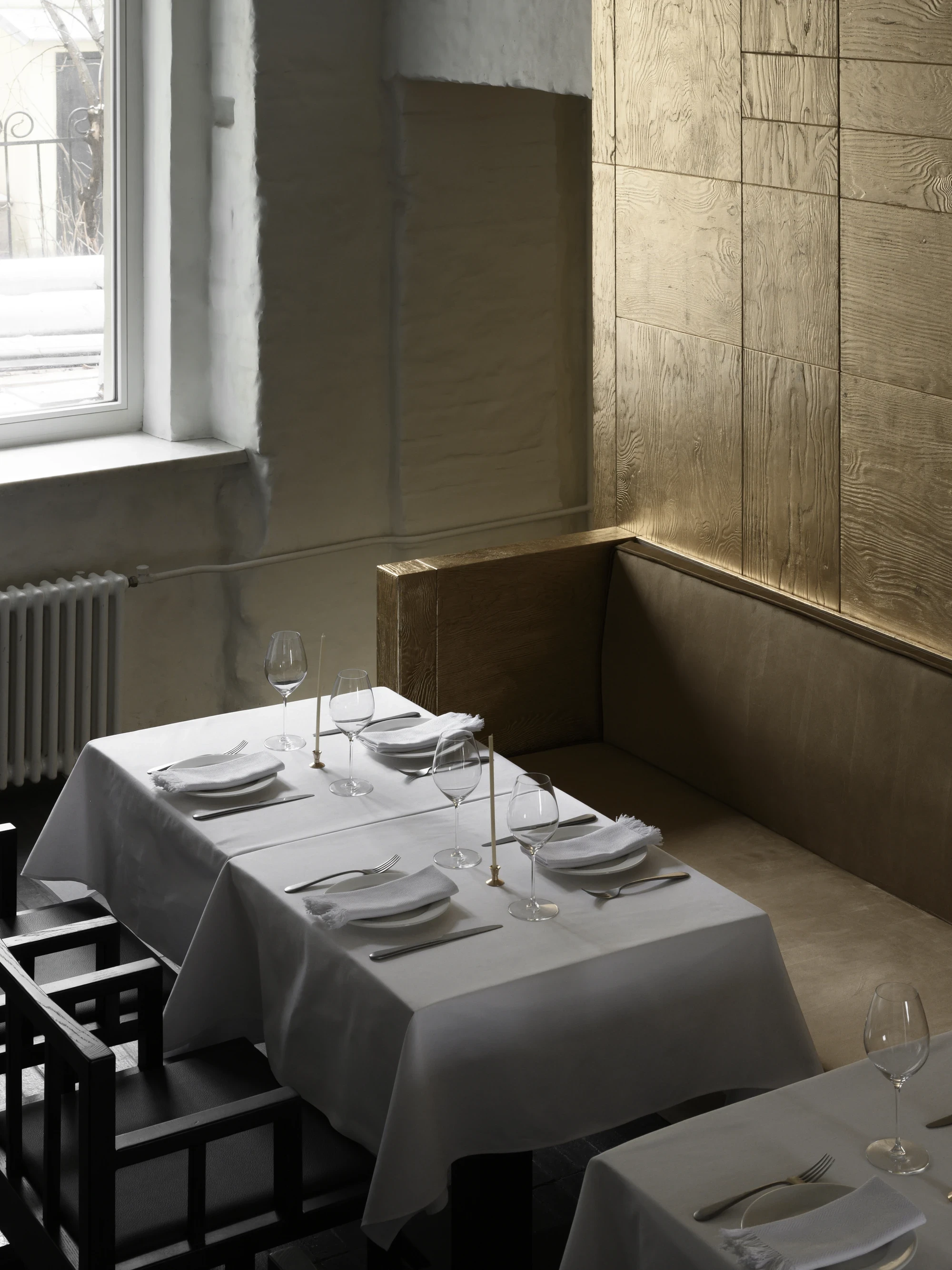 OKN Restaurant by SAGA, photographed by Daniel Annenkov — Overhead view of a banquette corner in daylight, showing the junction between rough white plaster and gold-leaf timber panel wall. A long rectangular table is set with multiple place settings.