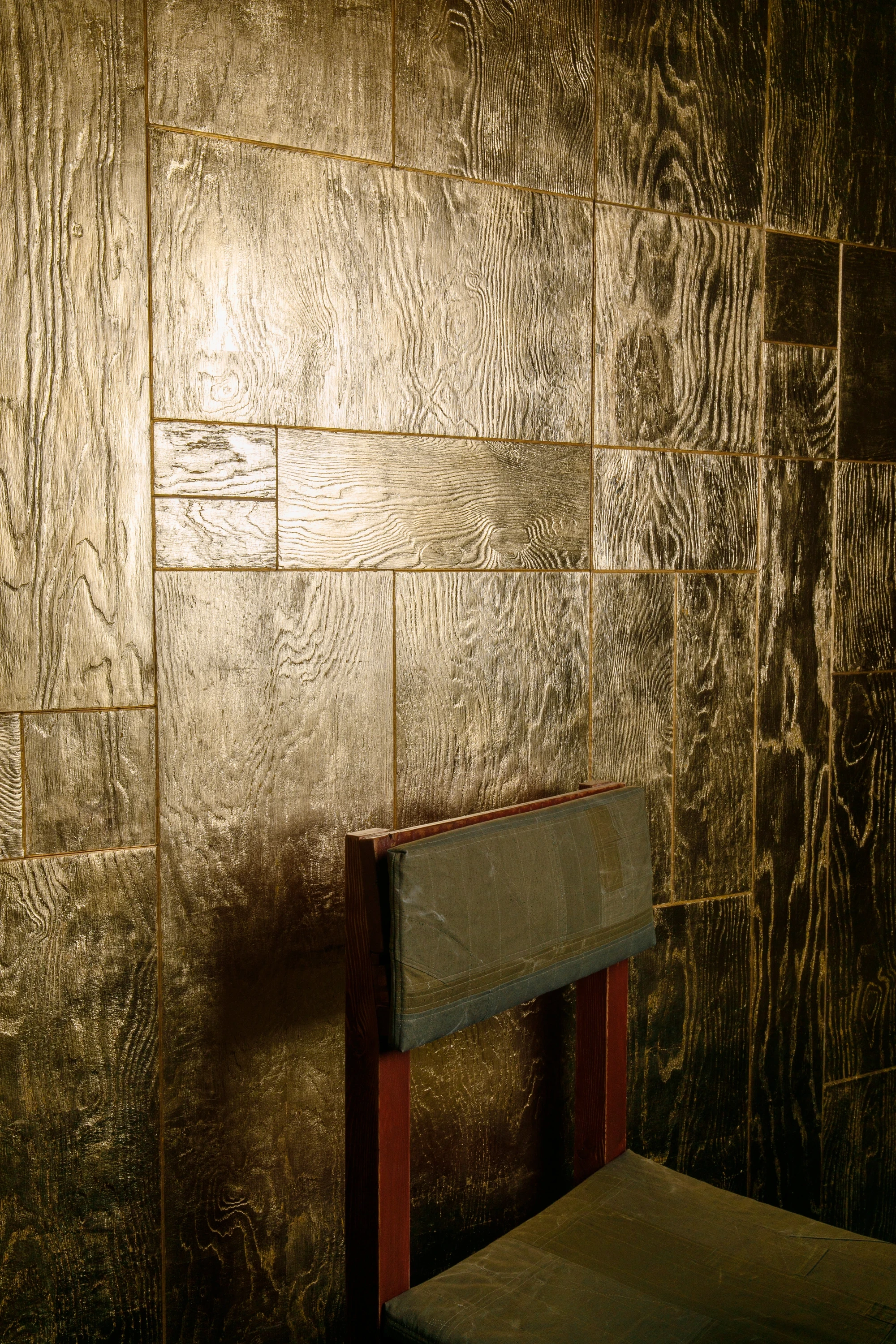 OKN Restaurant by SAGA, photographed by Daniel Annenkov — A single dining chair, its canvas-upholstered back set against the gold-leaf timber wall panels whose surface shows the transferred grain of the timber substrate.