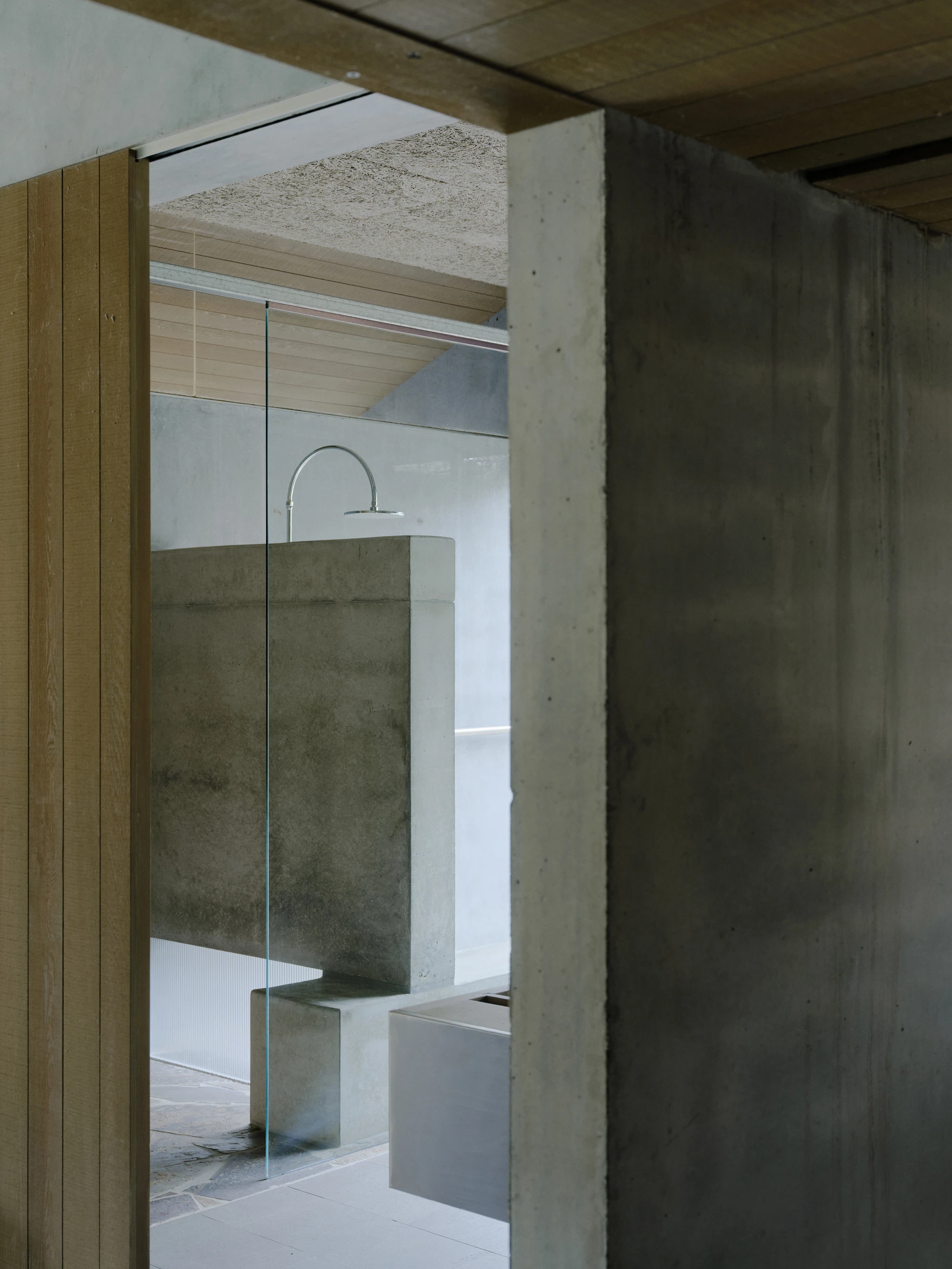This is Lumeah by Pepper & Well, photographed by Tom Ross — A framed glimpse into the ensuite seen between an existing timber-veneered cabinet door and a precast concrete column, the Stramit board ceiling transitioning from wood battens to fibrous board a