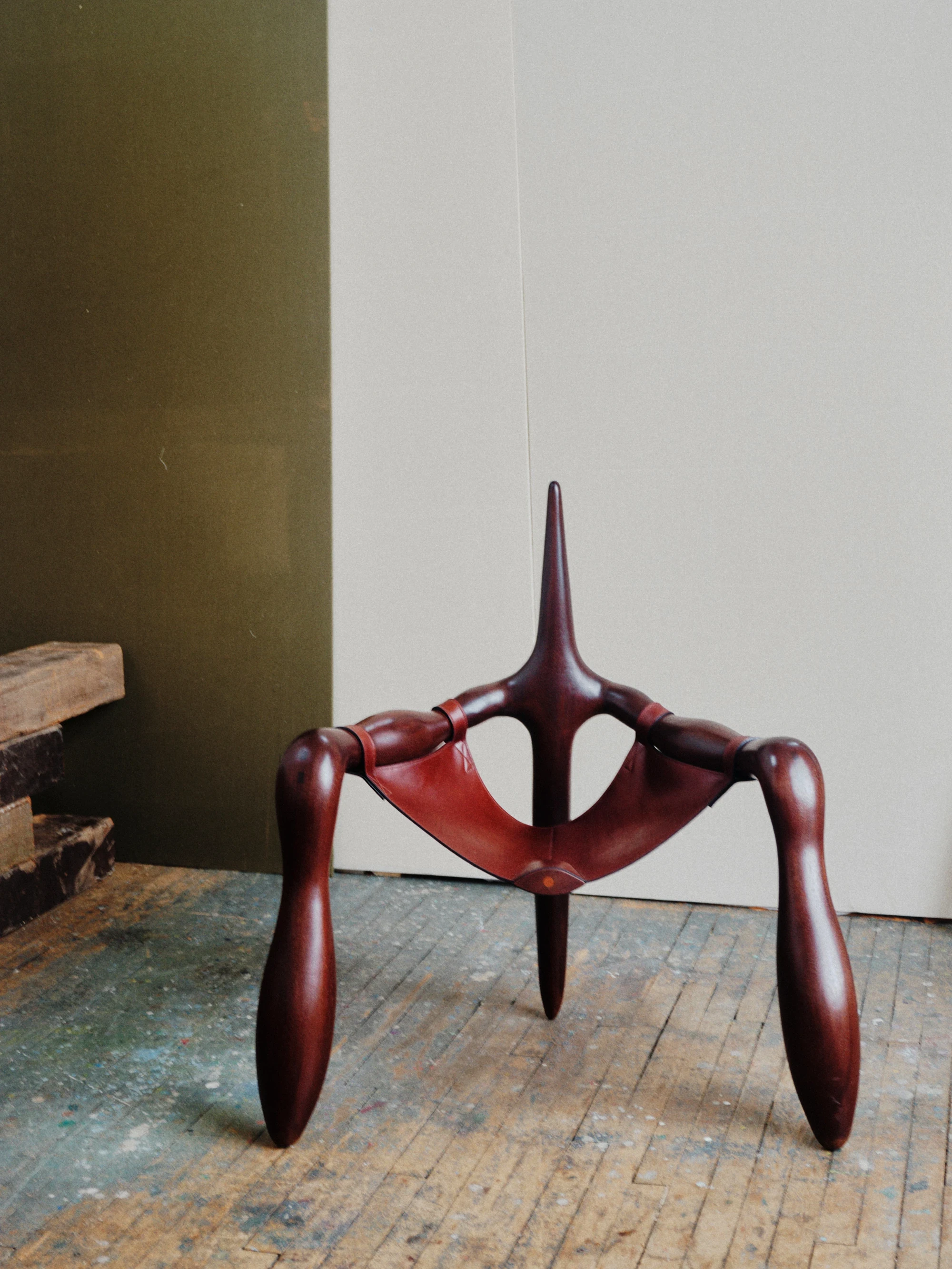 Instructions by Alberto Vitelio, photographed by @_victorjacques — the hand-carved Doussié chair stands against olive-green and off-white studio walls, its three bulbous legs on a paint-spattered floor, a narrow thorn-like spine rising from the oxblood fr