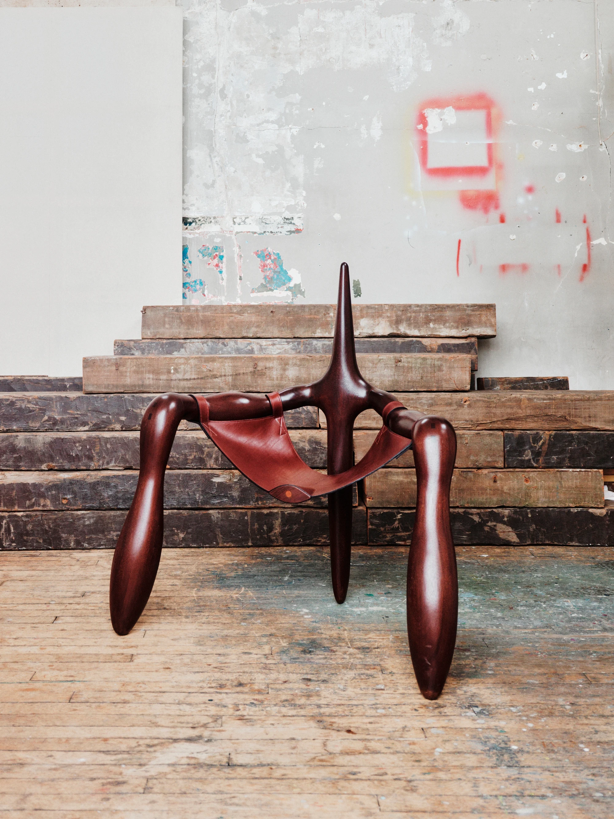 Instructions by Alberto Vitelio, photographed by @_victorjacques — the finished chair, oxblood Doussié and russet leather, photographed against the same distressed studio wall, the suspended seat visible in full from the front.