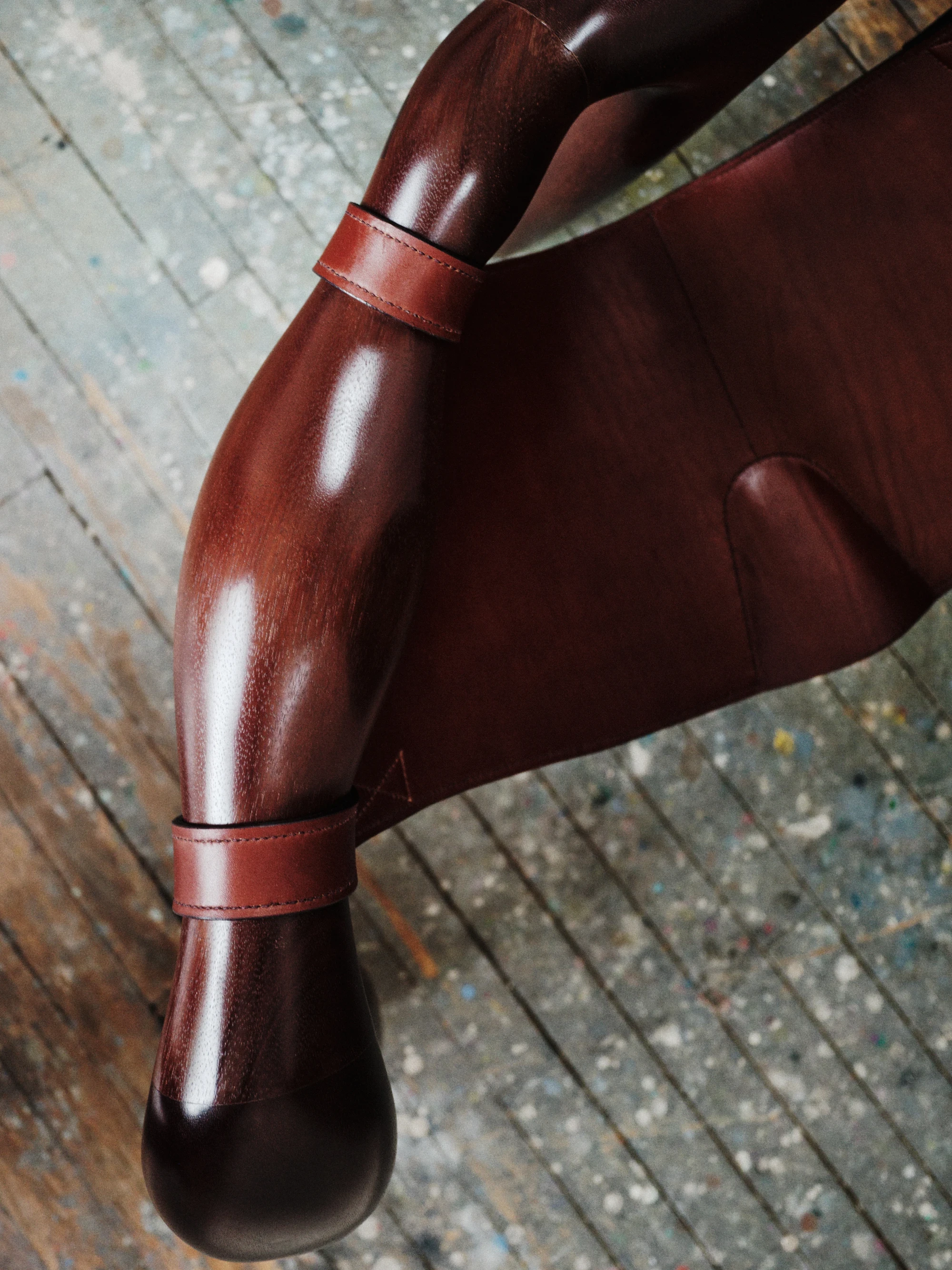 Instructions by Alberto Vitelio, photographed by @_victorjacques — a close detail of two hand-stitched leather straps cinching the junction of a Doussié leg to the seat rail, the polished wood grain visible against the paint-flecked studio floor.