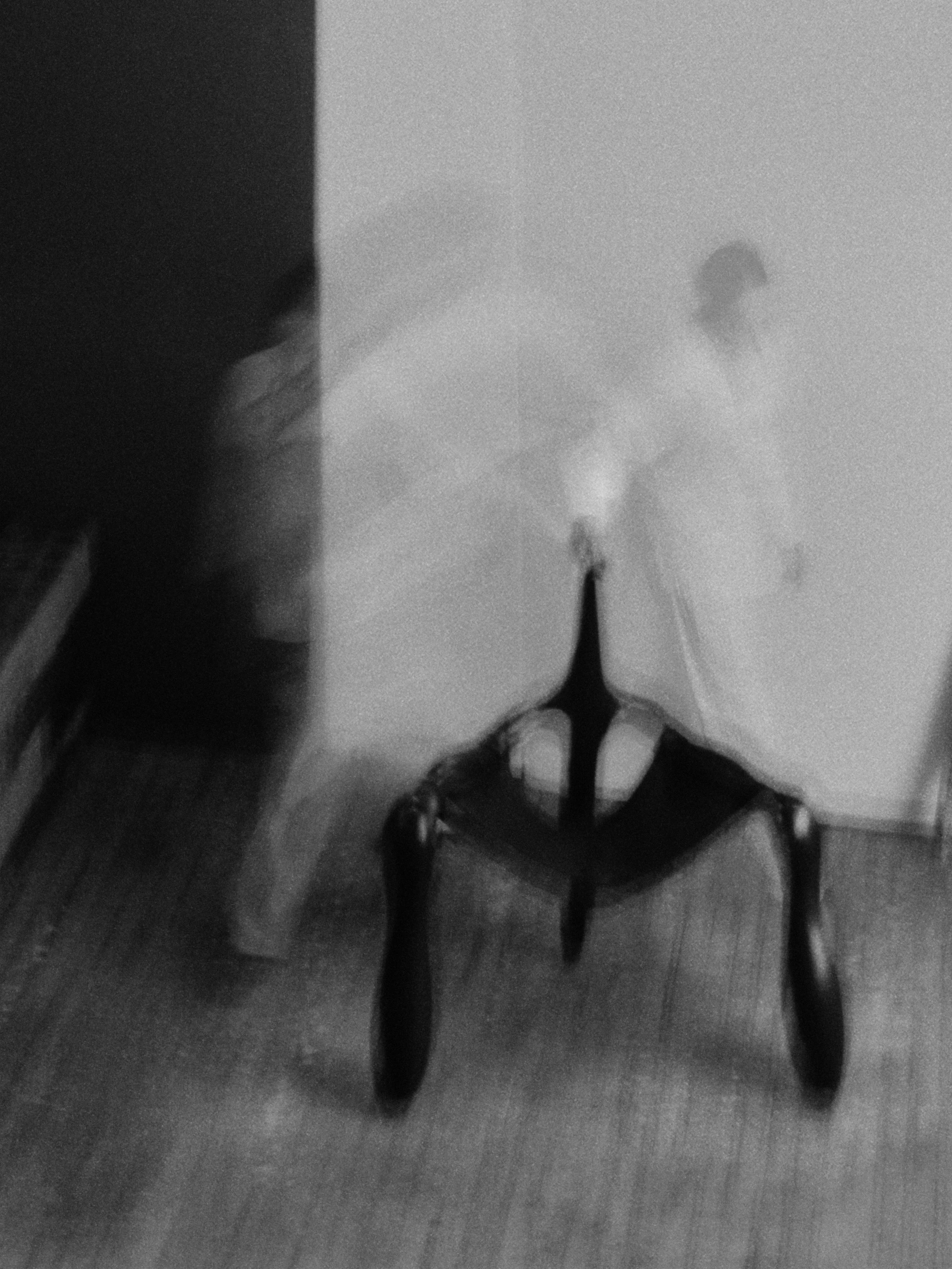 Instructions by Alberto Vitelio, photographed by @tautoism — a motion-blurred black-and-white image of a figure in a white garment moving with the chair, the long exposure collapsing the body and the object into a single indistinct form.