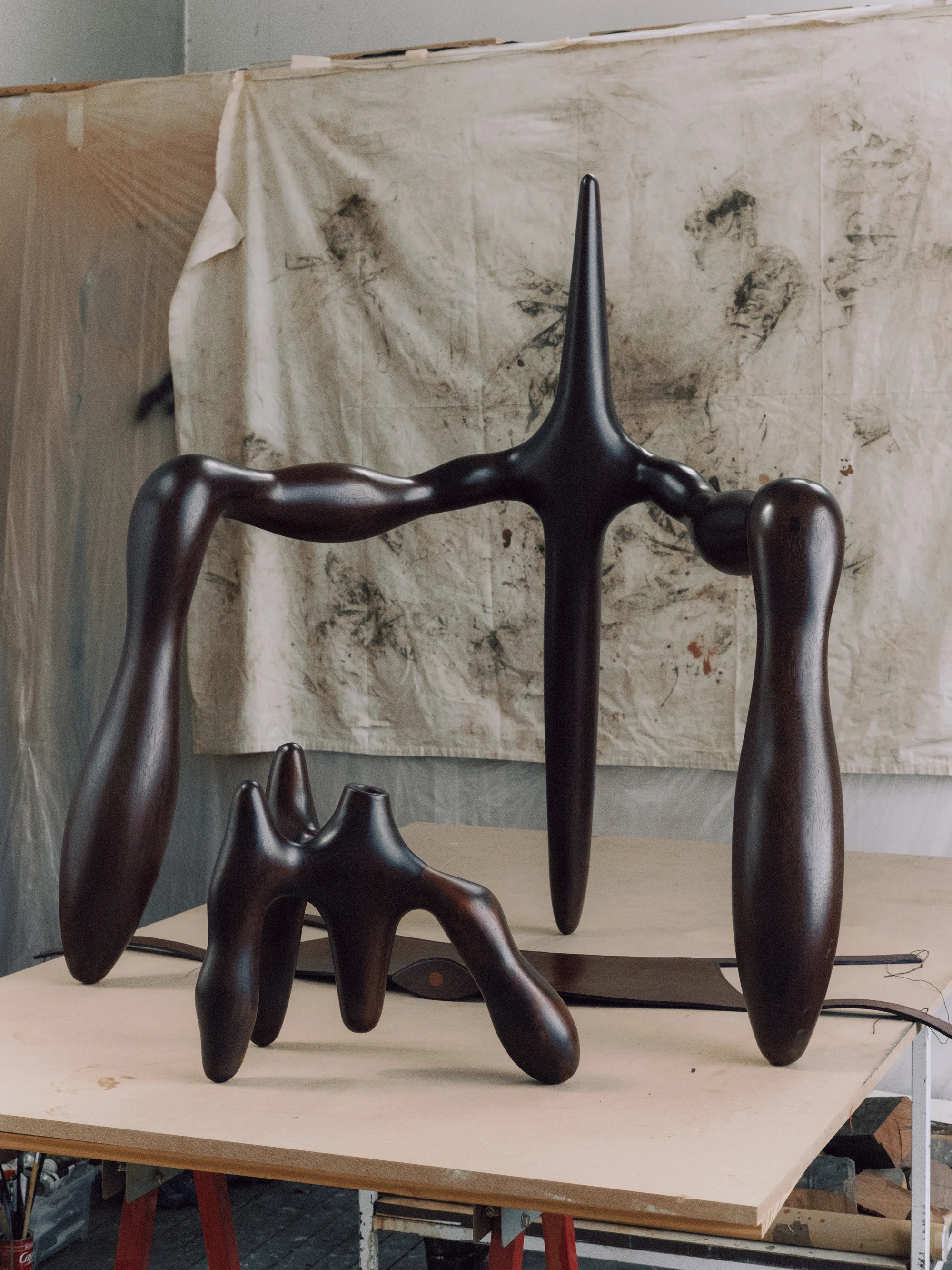 Instructions by Alberto Vitelio, photographed by @_victorjacques — a near-black Doussié armature and a small-scale maquette rest on a worktable in front of a canvas hung with charcoal figure drawings, revealing the sculptural process behind the finished c