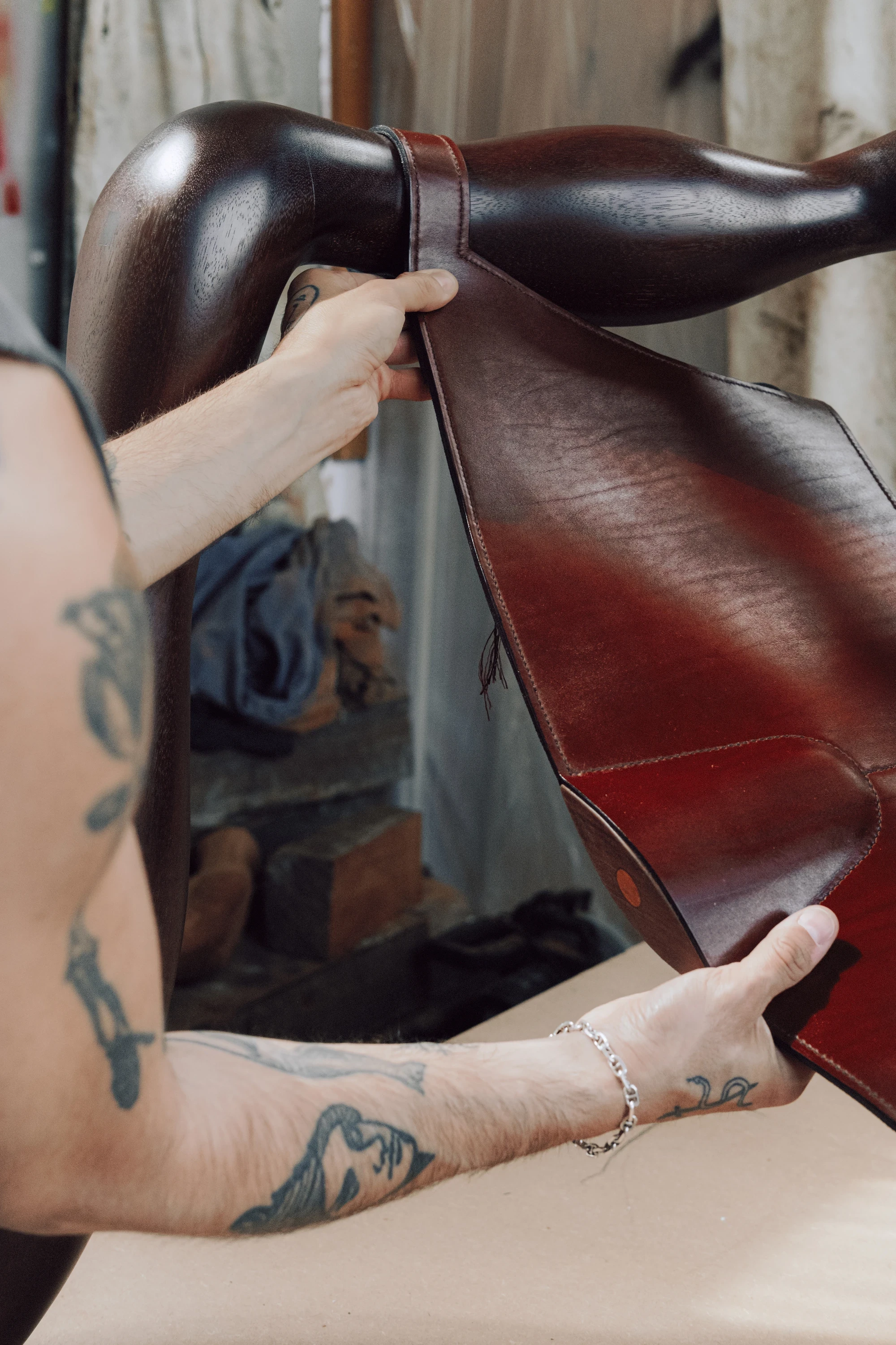 Instructions by Alberto Vitelio, photographed by @tautoism — tattooed arms lift the leather seat panel away from the Doussié frame to check its tension, the underside revealing hand stitching and a wooden corner plug, studio mirror reflected behind.