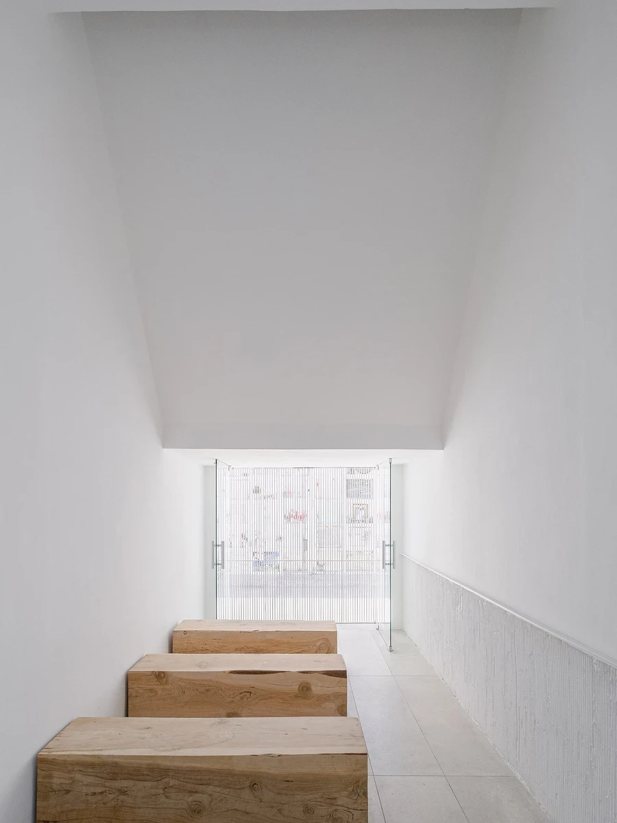 PSJ Chapel by Esteras Perrote Studio, photographed by Andrés Domínguez — raw live-edge timber steps descend inside the white chapel toward reeded cast-glass doors, the fine-ribbed render wainscot visible on the right wall under a tall splayed ceiling volu