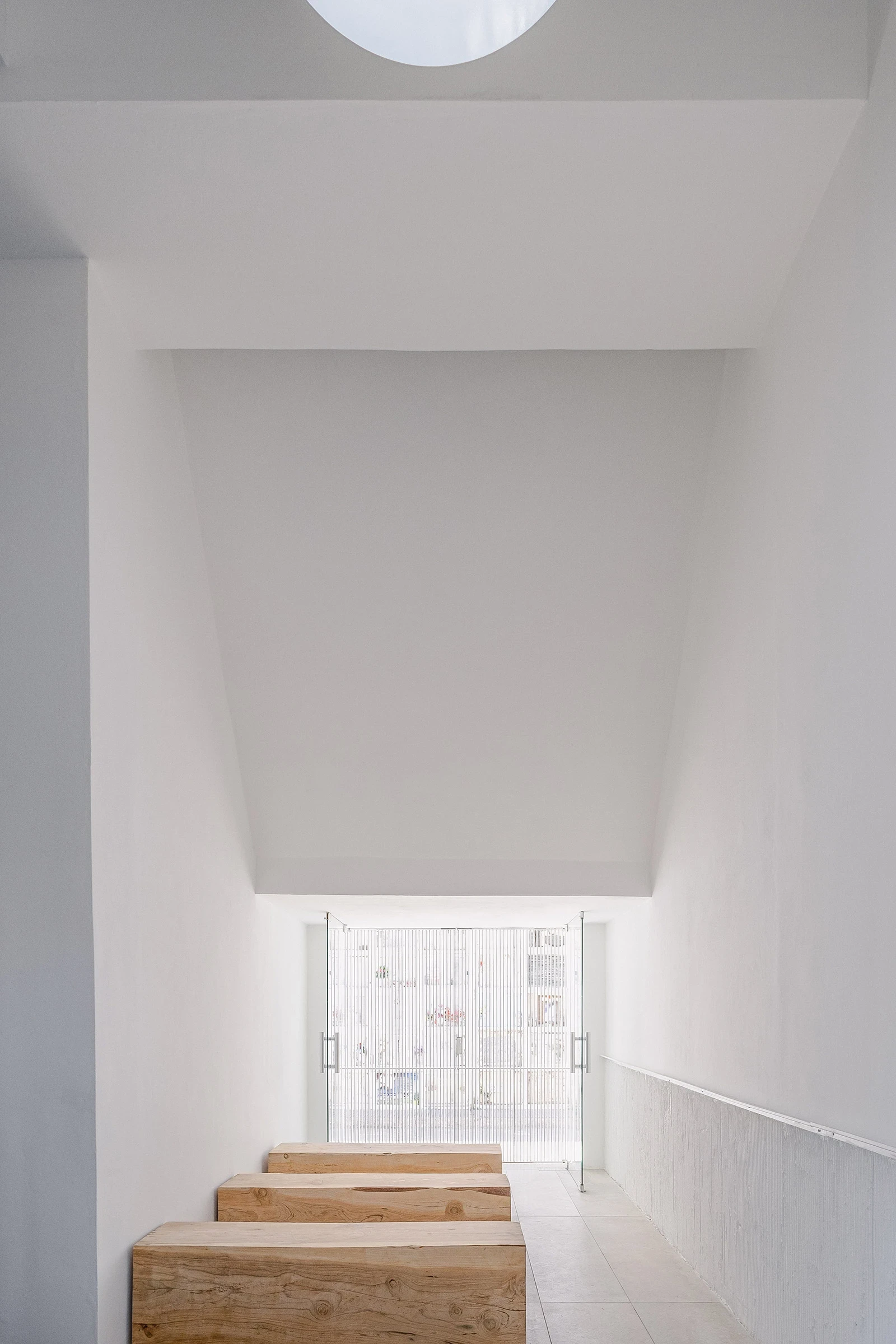 PSJ Chapel by Esteras Perrote Studio, photographed by Andrés Domínguez — interior looking toward the street, the circular ceiling oculus partially visible upper left, raw timber slabs in the midground, reeded glass doors transmitting a blurred exterior vi