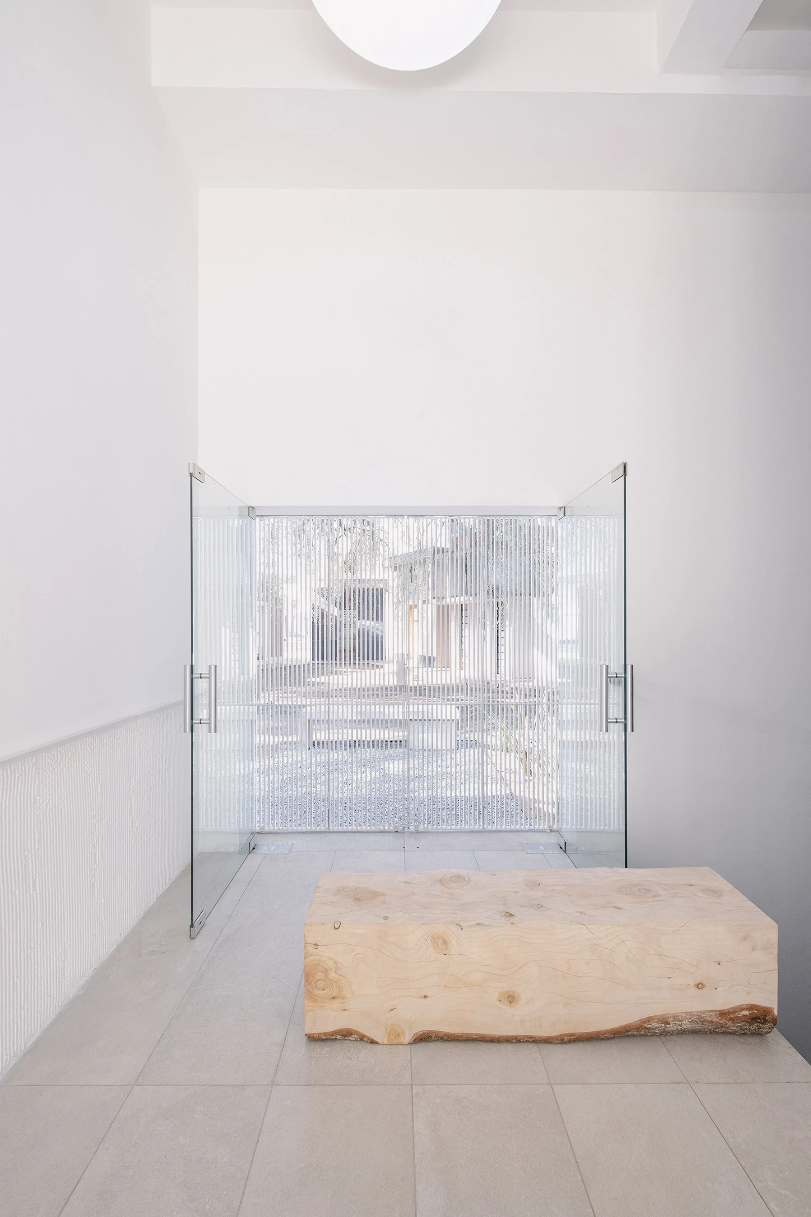 PSJ Chapel by Esteras Perrote Studio, photographed by Andrés Domínguez — a single live-edge timber slab rests on the large-format grey tile floor, bark edge intact, the reeded glass double door open behind it and the ribbed render wainscot on the left wal