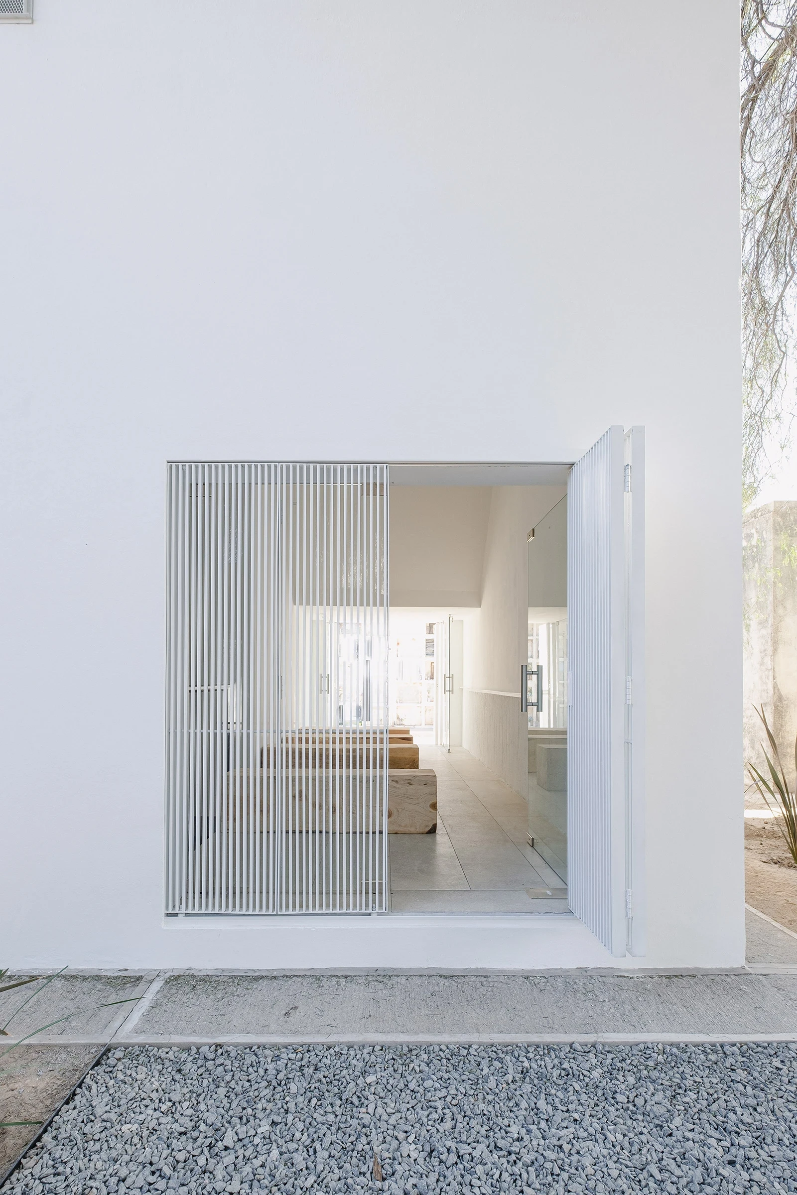PSJ Chapel by Esteras Perrote Studio, photographed by Andrés Domínguez — the white louvred panel folded back against the portal reveal, the reeded glass door beyond partially open, grey gravel path in the foreground and the dry cemetery ground extending l