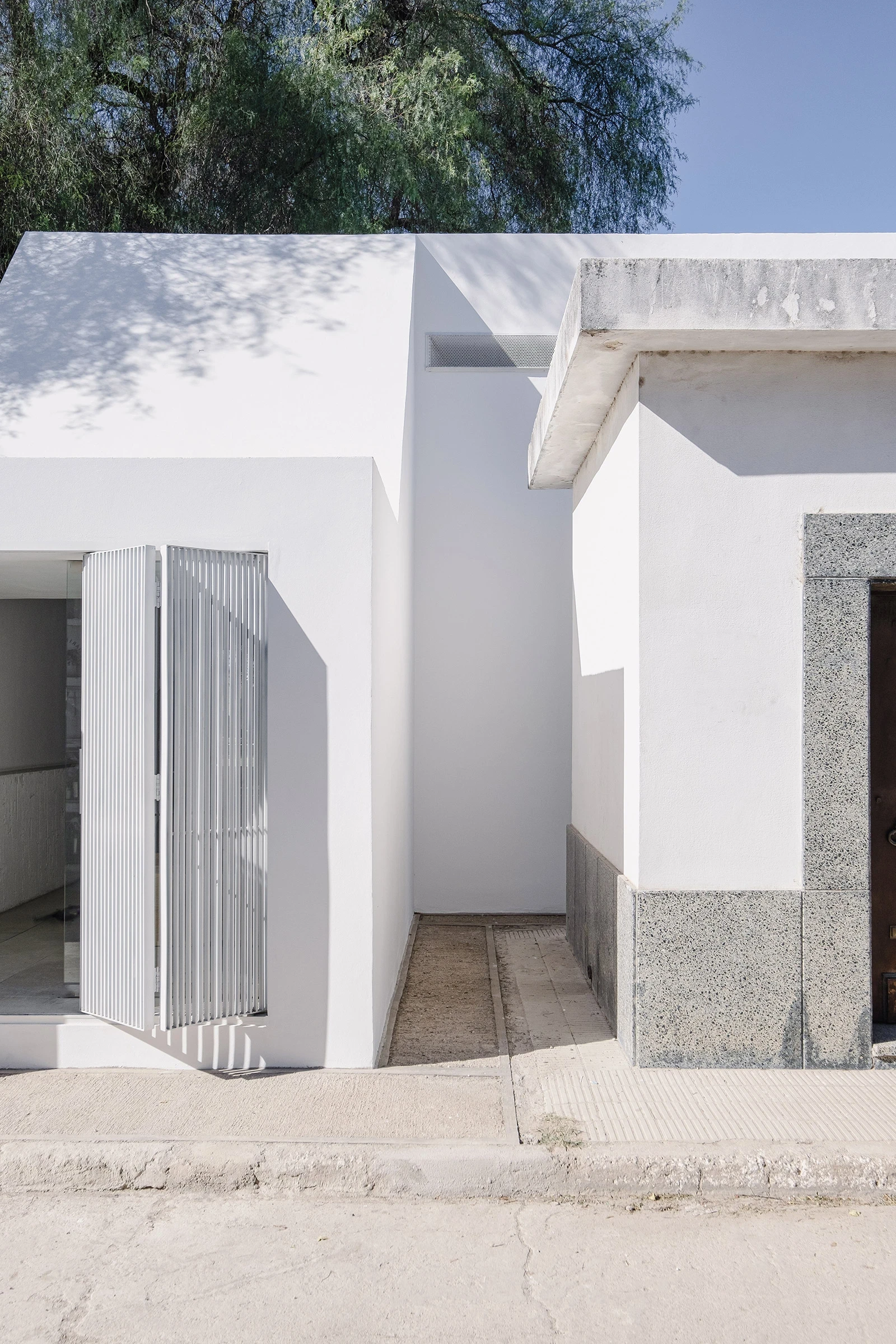 PSJ Chapel by Esteras Perrote Studio, photographed by Andrés Domínguez — street-facing façade in full sun, white rendered planes intersecting at a sharp oblique angle, with a louvred aluminium gate on the left face and a ribbed stone-aggregate panel along