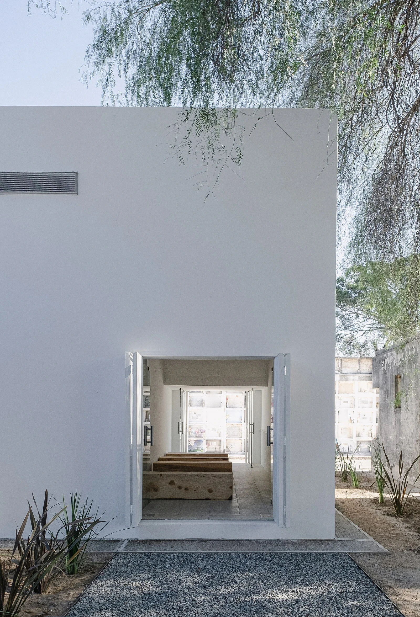 PSJ Chapel by Esteras Perrote Studio, photographed by Andrés Domínguez — straight shot through the open square portal, two live-edge timber slabs visible inside, the reeded glass doors at the far end transmitting light from the street beyond.