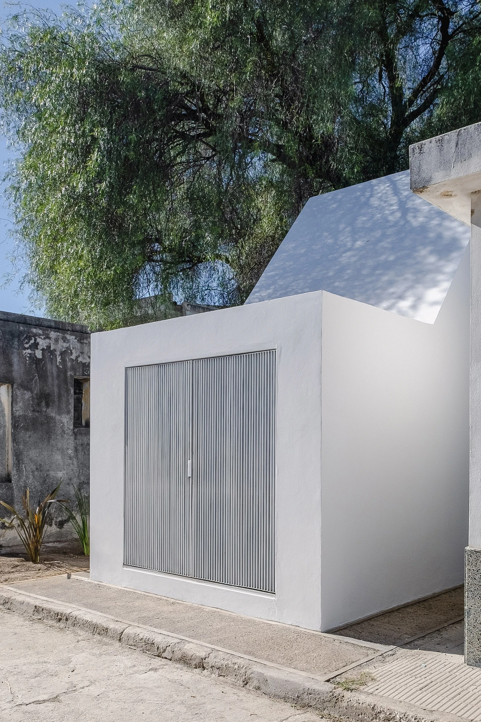 PSJ Chapel by Esteras Perrote Studio, photographed by Andrés Domínguez — street-facing volume from the pavement, full-width ribbed corrugated metal doors set into white render, the angled roof plane above and the pepper tree overhanging the parapet.