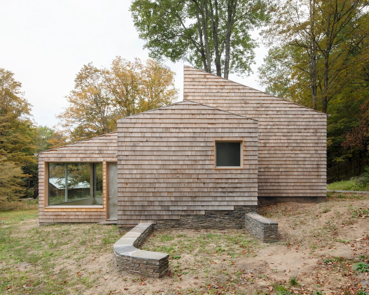 Studio & Guest House by Neil Logan, photographed by Mikael Olsson — rear elevation of the guest house, cedar shingles wrapping the full volume, a large square picture window set with a fir reveal, dry-stack stone steps at grade.