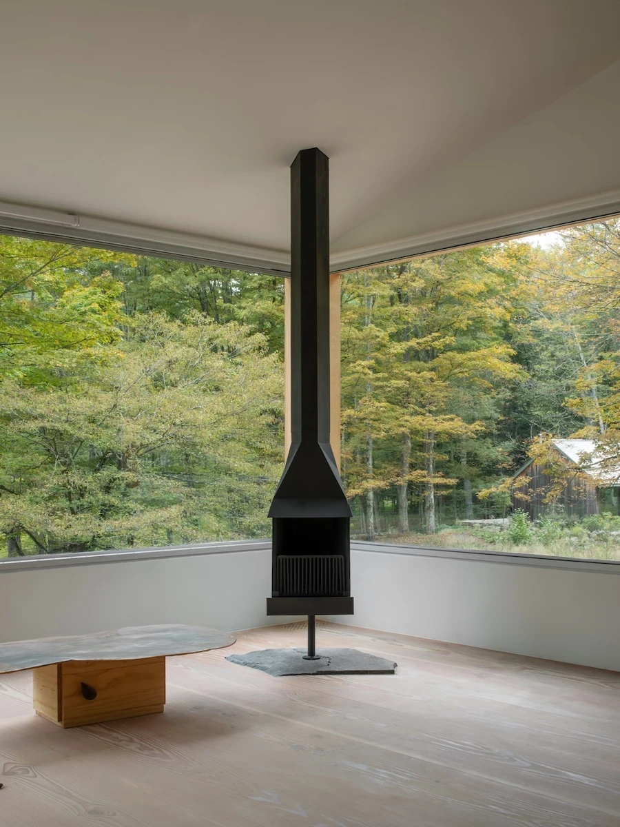 Studio & Guest House by Neil Logan, photographed by Mikael Olsson — corner panoramic glazing wraps two walls of the guest house interior, framing the forest; a black suspended wood-burning stove stands on a slate hearth over wide-plank pale fir floor.