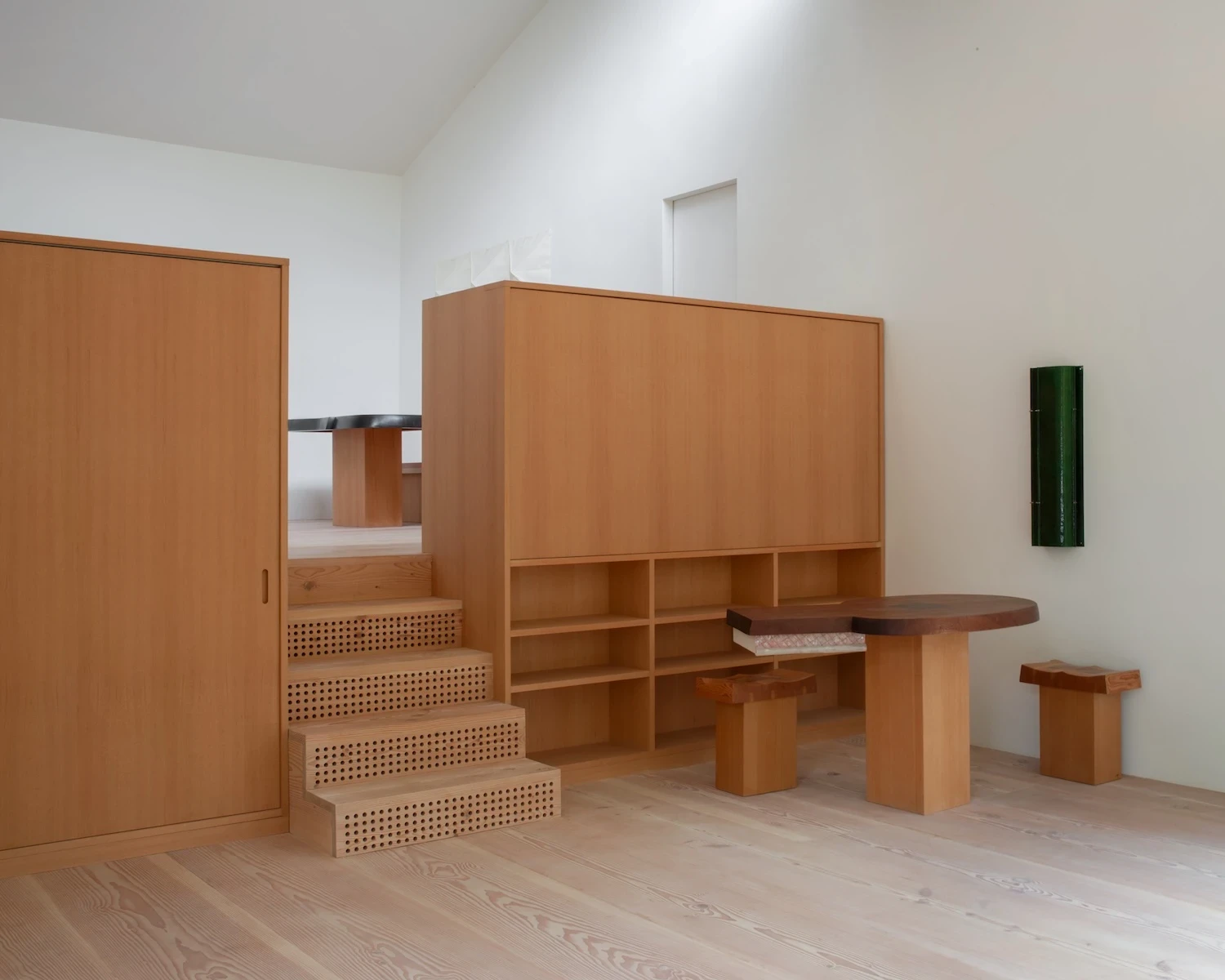 Studio & Guest House by Neil Logan, photographed by Mikael Olsson — cane-front stair risers climb past open fir shelving to the upper level; the dark walnut table sits at the base of the unit, a folded canvas textile hanging on the white plaster wall behi