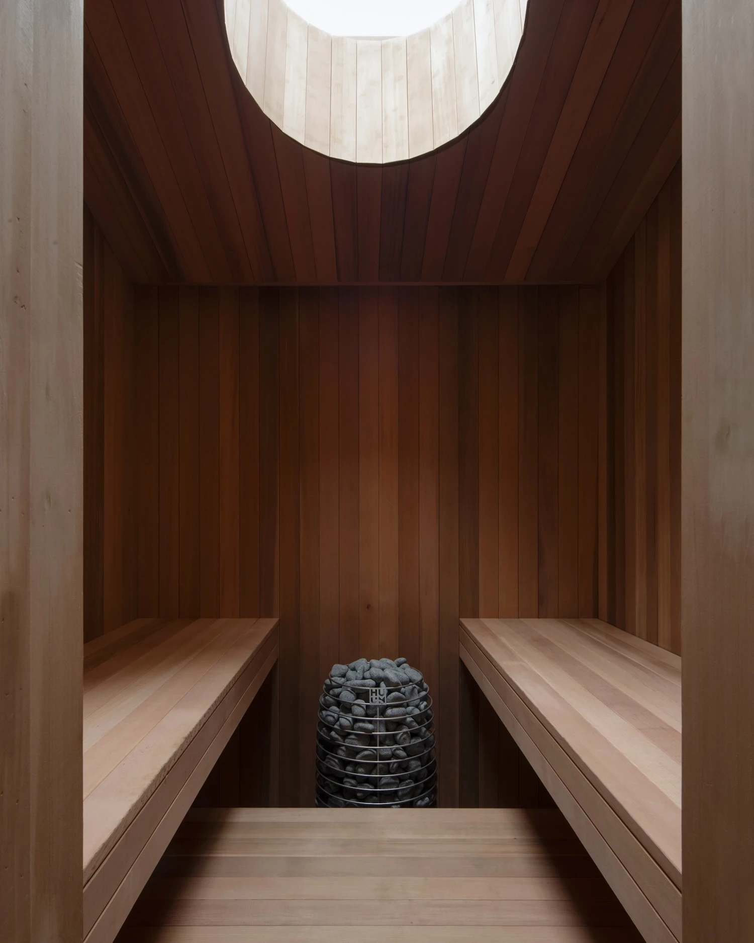 Studio & Guest House by Neil Logan, photographed by Mikael Olsson — cedar sauna interior with bench seating on both sides, a Huum electric stone heater at the far end, and a circular light well cut into the barrel-vaulted cedar ceiling above.