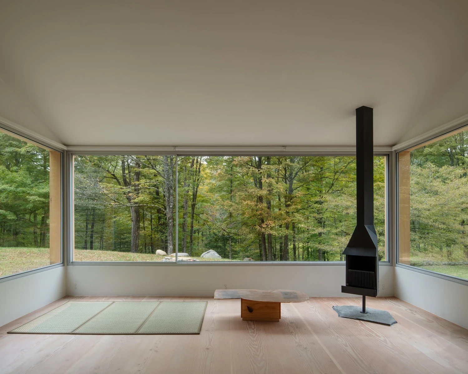 Studio & Guest House by Neil Logan, photographed by Mikael Olsson — guest house interior with tatami mat on fir floor, the panoramic glazing wrapping the corner to the tree line, and the black suspended stove visible to the right on its slate hearth.
