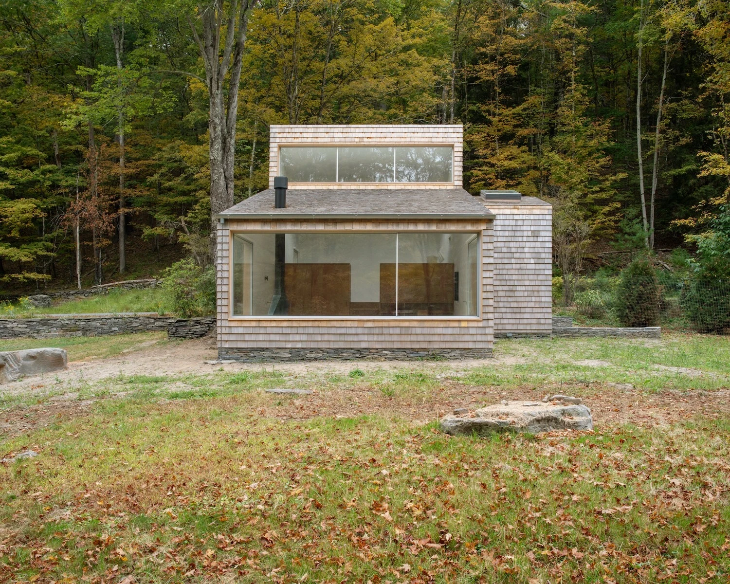 Studio & Guest House by Neil Logan, photographed by Mikael Olsson — guest house exterior facing the clearing: cedar shingle cladding, a clerestory band running above the main glazed opening, and a small chimney stack above the roofline.