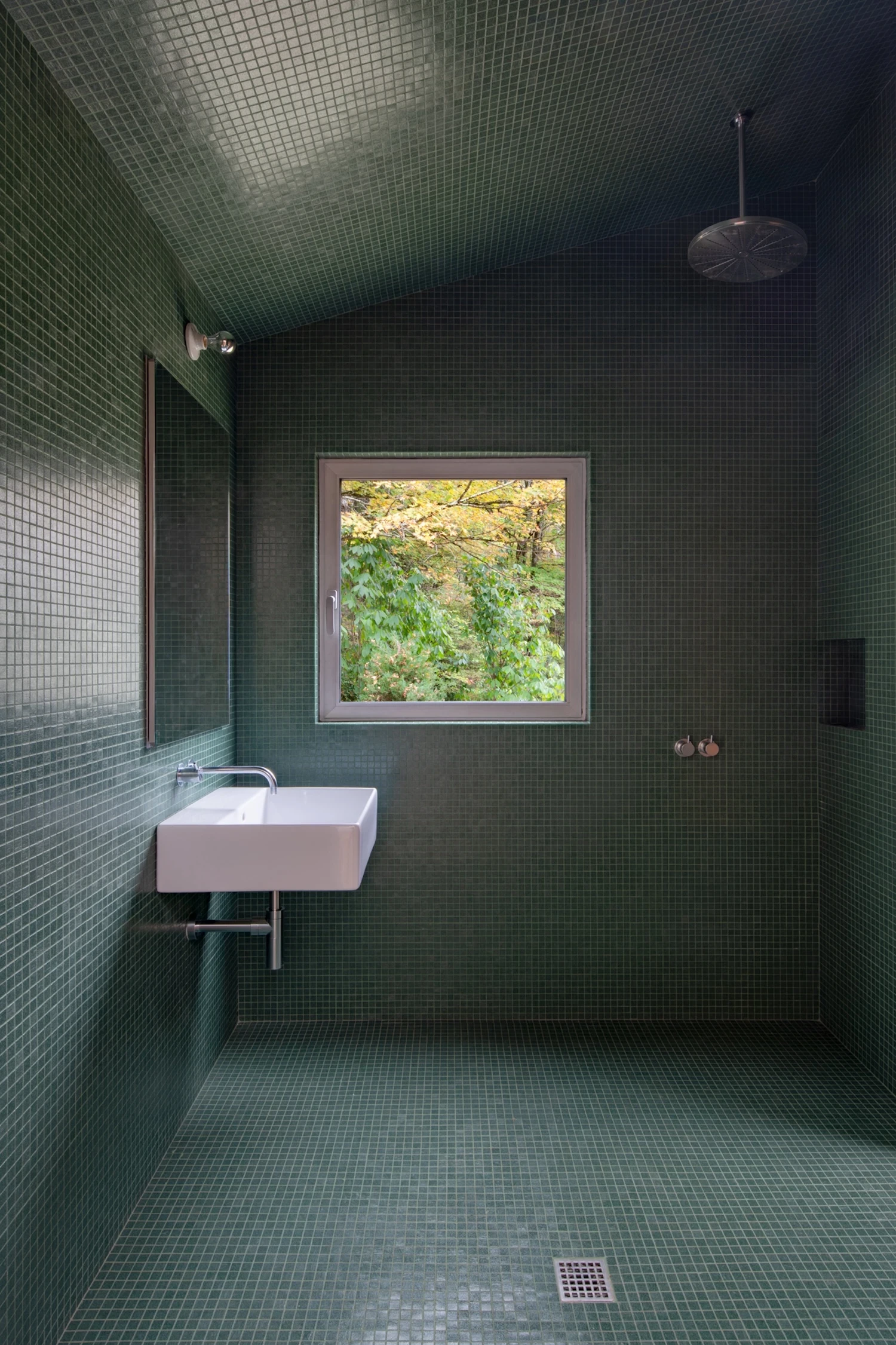 Studio & Guest House by Neil Logan, photographed by Mikael Olsson — guest house bathroom tiled floor-to-ceiling in deep forest-green small mosaic, a square window framing autumn foliage, wall-mounted ceramic sink, and an overhead rain showerhead.