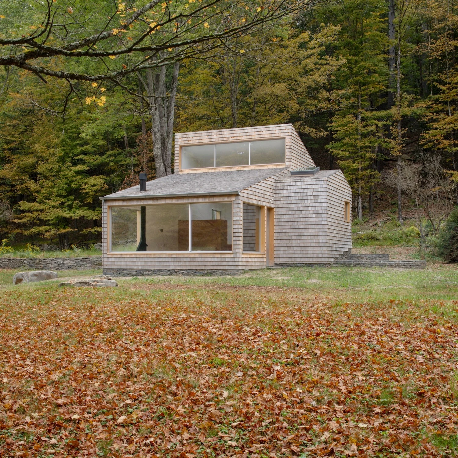 Studio & Guest House by Neil Logan, photographed by Mikael Olsson — guest house from across the meadow in full autumn leaf fall, cedar shingles weathering to grey-gold, the stovepipe chimney rising above the ridgeline against deciduous forest.