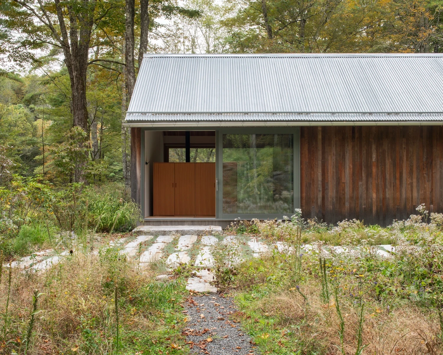 Studio & Guest House by Neil Logan, photographed by Mikael Olsson — studio approach path of gravel and stone pavers leading to a warm fir entry door set within weathered dark vertical board cladding, corrugated metal roof above, wildflowers at the verge.
