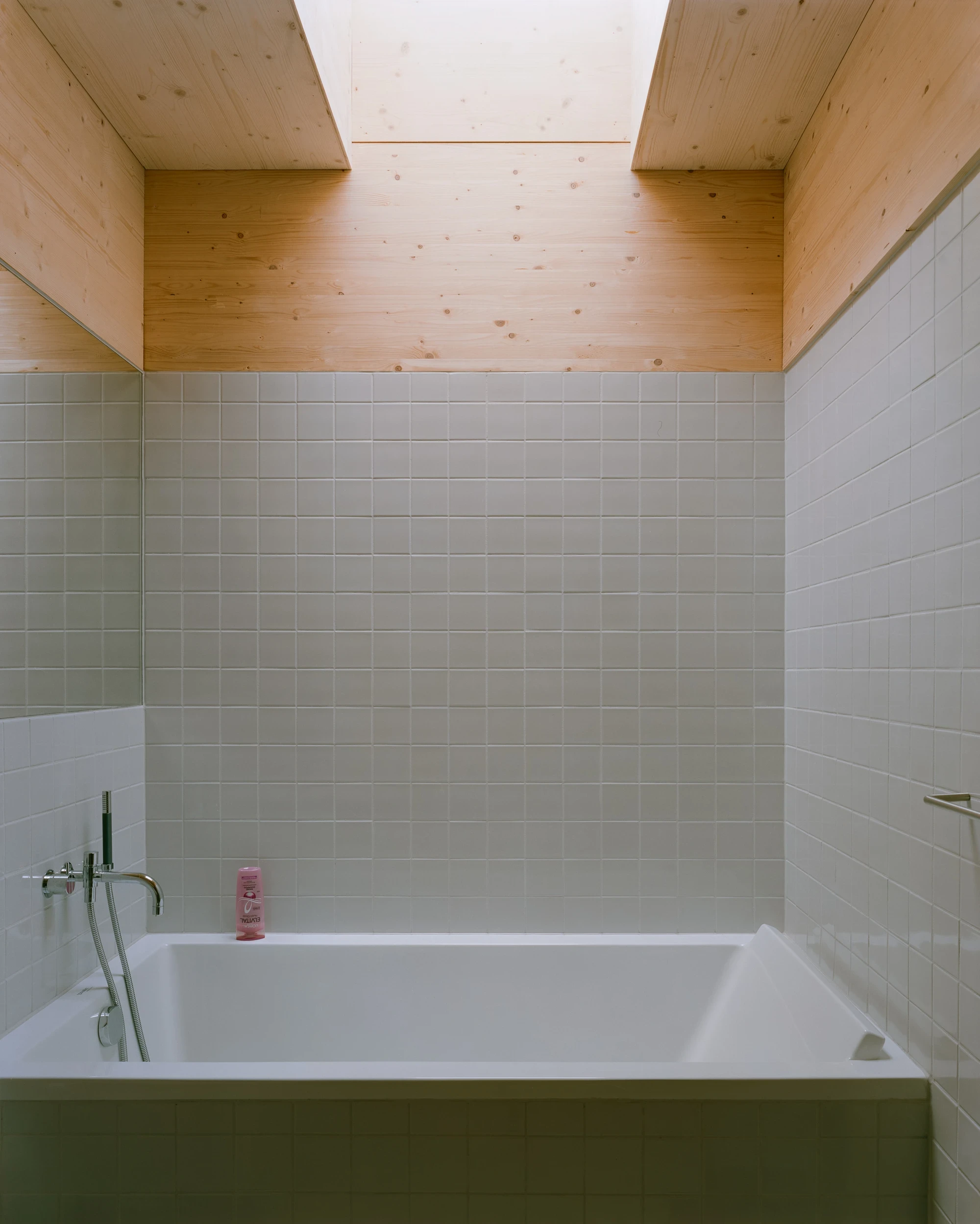 House in Ebeltoft by Høyer Arkitektur, photographed by Hampus Berndtson — the bathroom, square off-white ceramic tile running to mid-wall height with spruce CLT above, a ceiling rooflight, and a chrome wall-mounted bath tap.