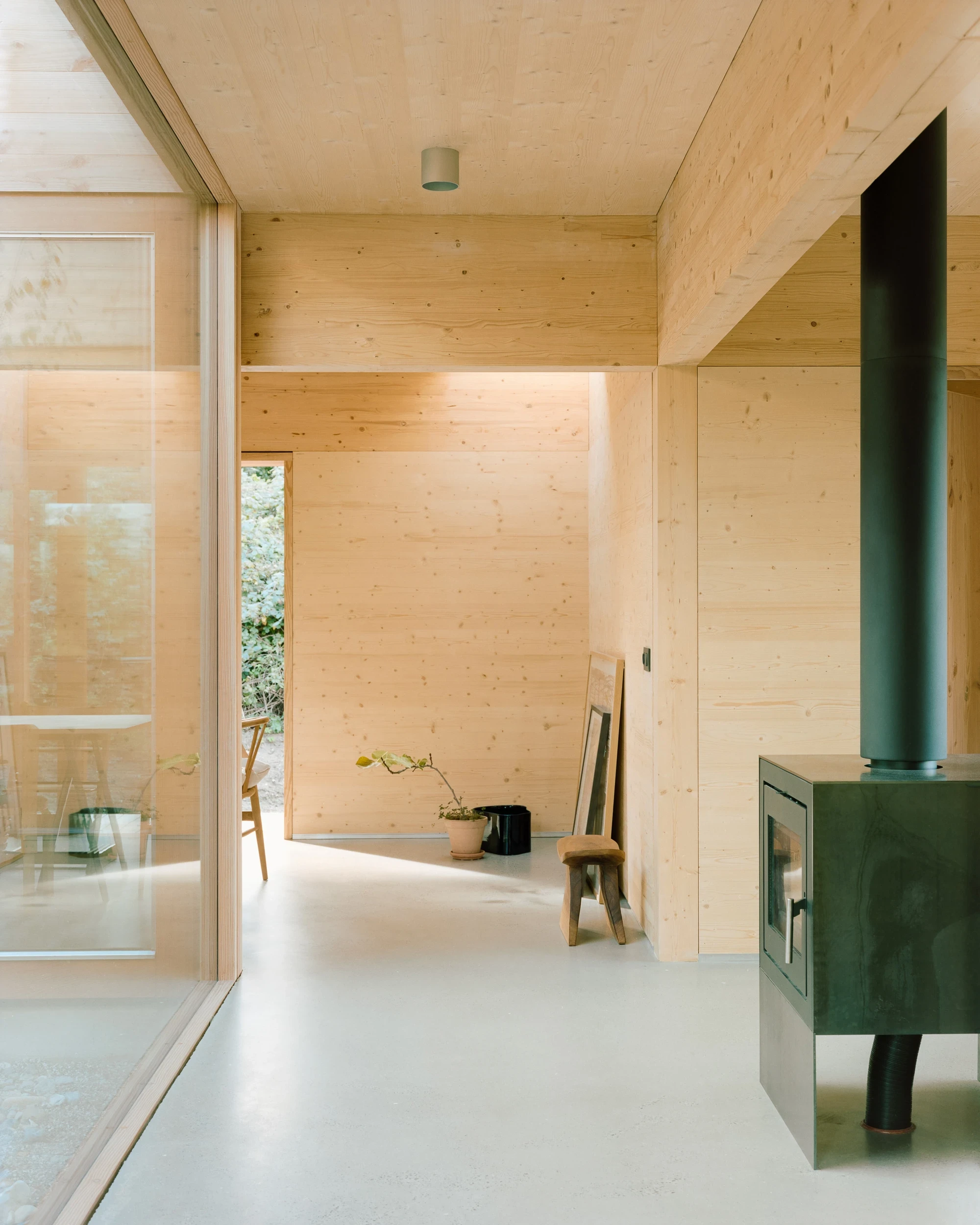 House in Ebeltoft by Høyer Arkitektur, photographed by Hampus Berndtson — a secondary room with the dark olive-green cast-iron log stove and matching enamel flue pipe, CLT walls, and a view through an open partition to the atrium.