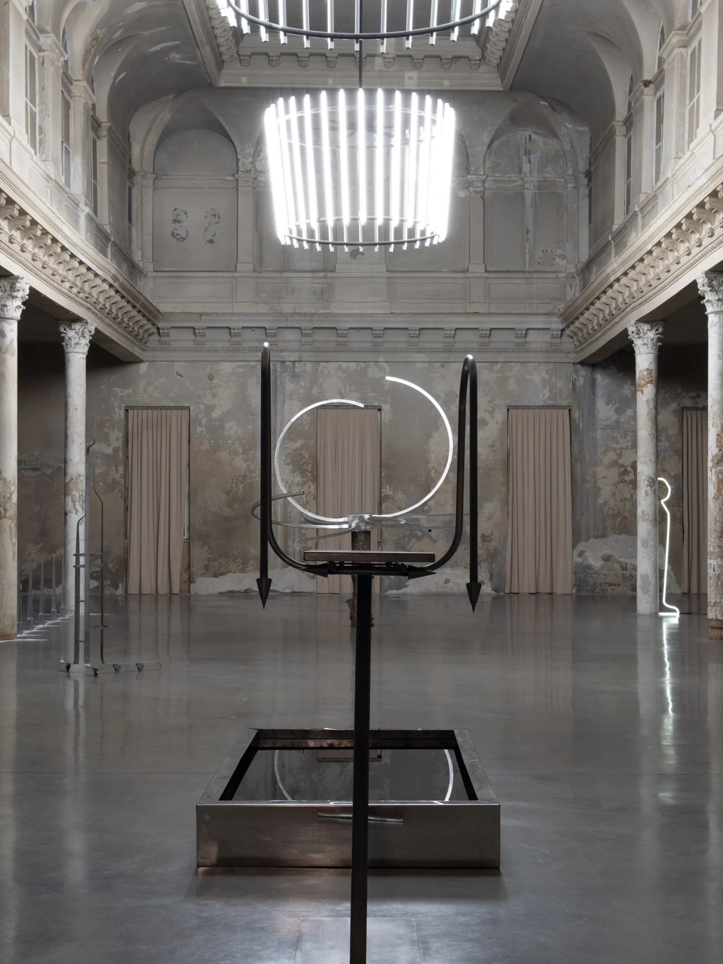 Everything Comes Together While Pushing All Apart by Roxana Morar — Wide install view with a tall steel framework by Claudiu Lazar centred under the suspended cage light, mirror trough at its base, Beaux-Arts hall stretching behind.
