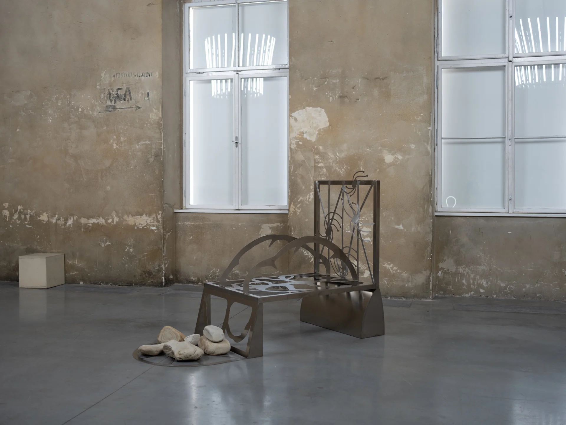 Everything Comes Together While Pushing All Apart by Roxana Morar — Stainless bench by Ana Petrovici with organic cut-outs and a small pile of river stones nesting underneath, set between two industrial windows.