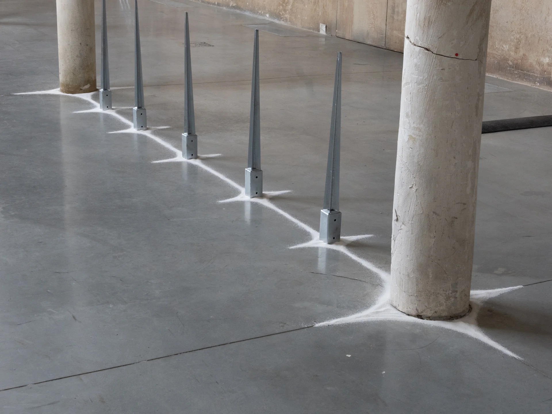 Everything Comes Together While Pushing All Apart by Roxana Morar — A row of six galvanized stakes by Ana Ionescu walking across the polished concrete floor, each anchored to a painted star and pointing at the ceiling.